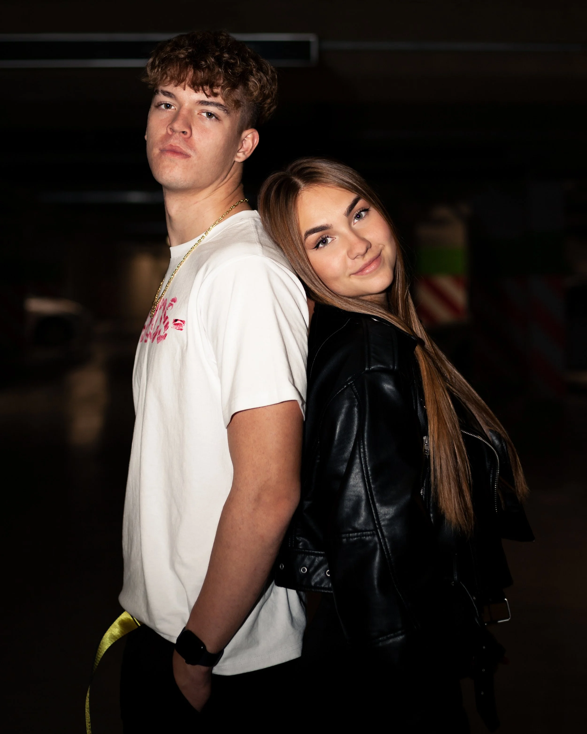 A young man with curly brown hair and a young woman with long brown hair pose back-to-back in a dark indoor setting. The man wears a white T-shirt with pink writing, a gold chain, and a black fitness tracker. The woman wears a black leather jacket and has a slight smile.