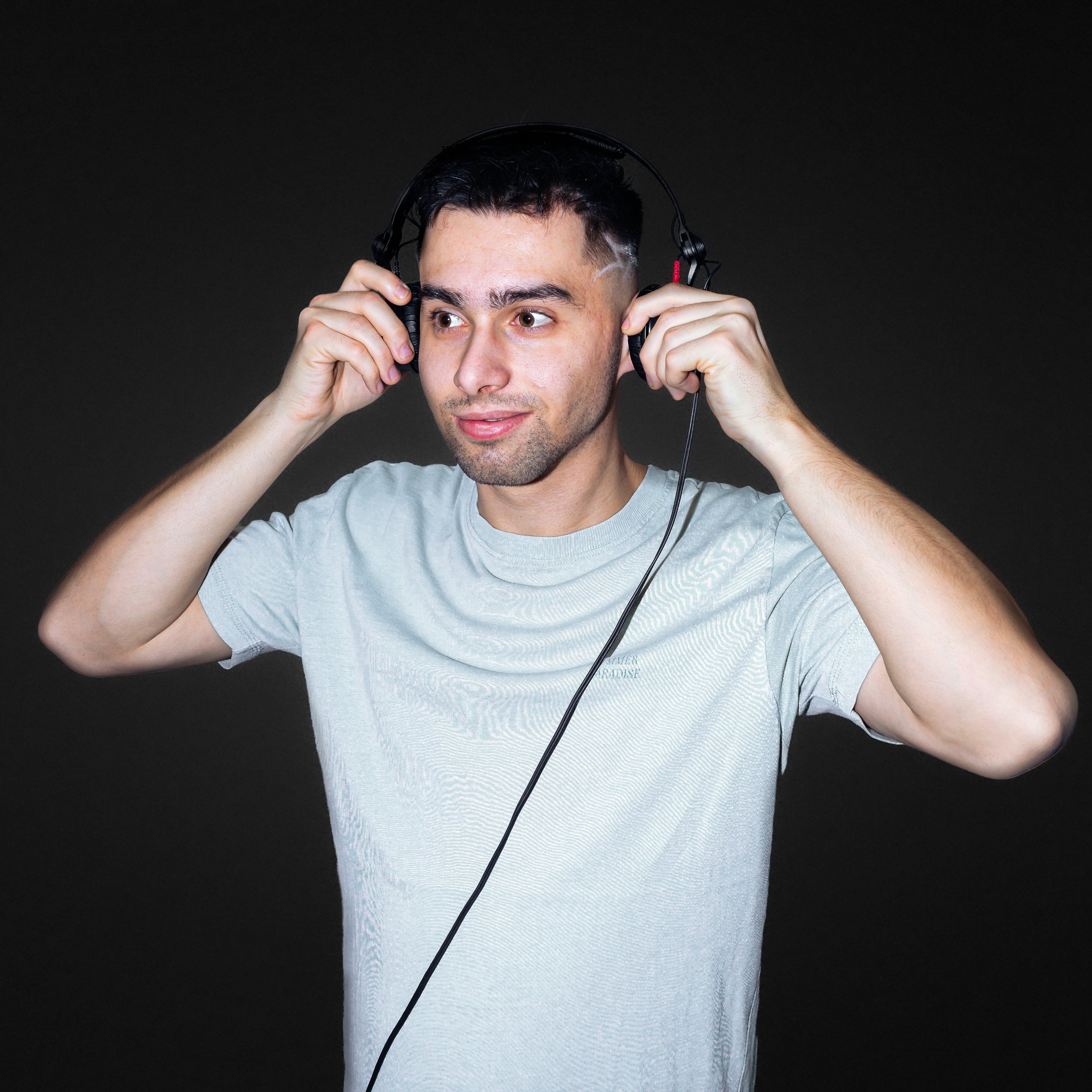 A young man with short dark hair, wearing a light gray T-shirt, is holding a pair of black over-ear headphones to his ears against a plain dark background, appearing to be adjusting or listening to the headphones.
