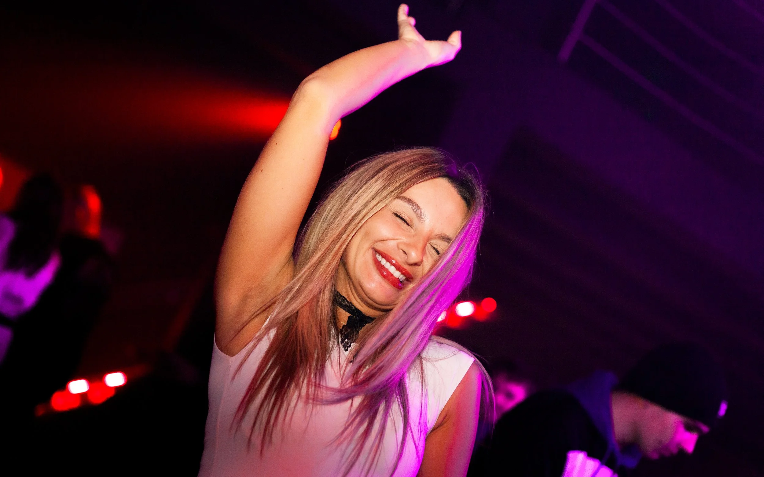 A young woman with long blonde hair dancing at a party with purple and red lighting, smiling with her eyes closed, wearing a sleeveless top and black lace choker.