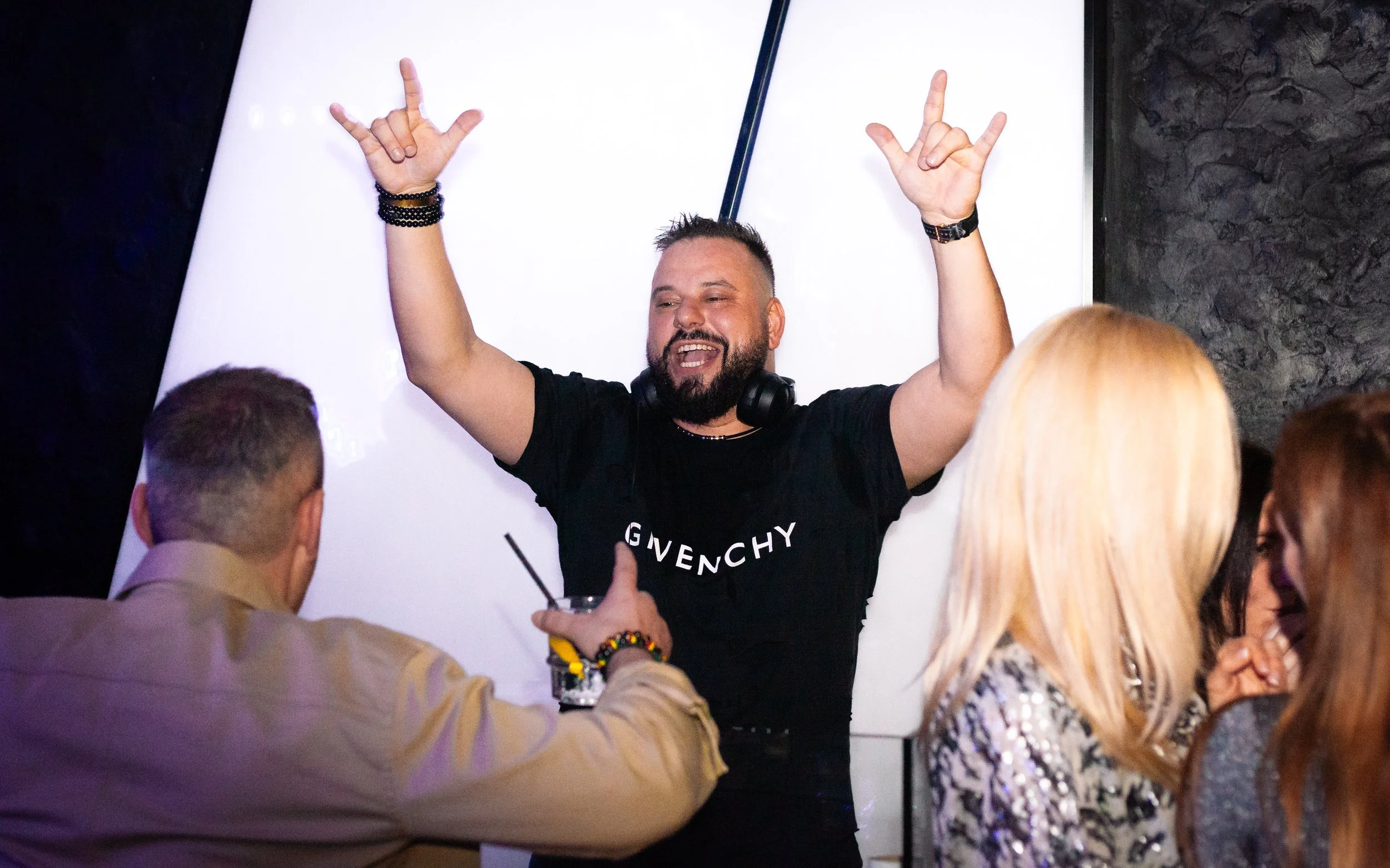 A man with a beard and short hair wearing a black Givenchy t-shirt and a black watch, raising his hands with rock-and-roll gesture, surrounded by a group of people, in a dimly lit setting.