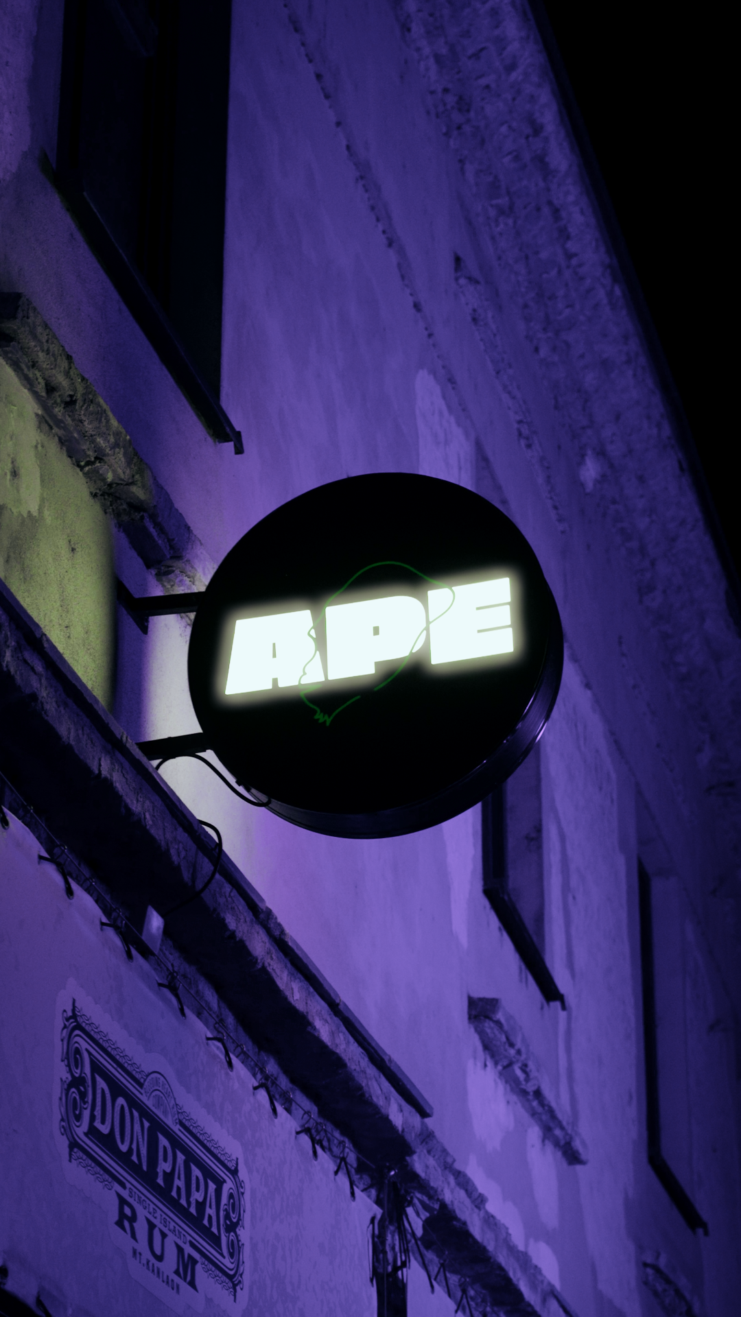 Neon sign with the word "APPE" glowing white, outlined by a green line, mounted on a textured wall with purple lighting and visible electrical cords.