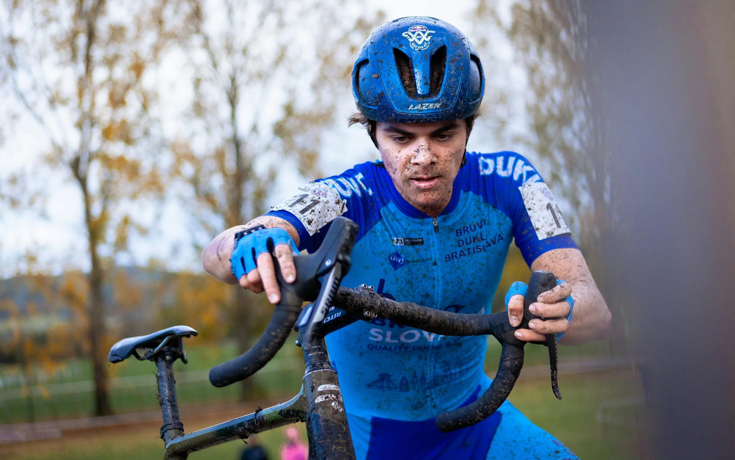 A male cyclist wearing a blue helmet and a matching blue cycling outfit, covered in mud, riding a mountain bike outdoors in a wooded area with autumn foliage in the background.