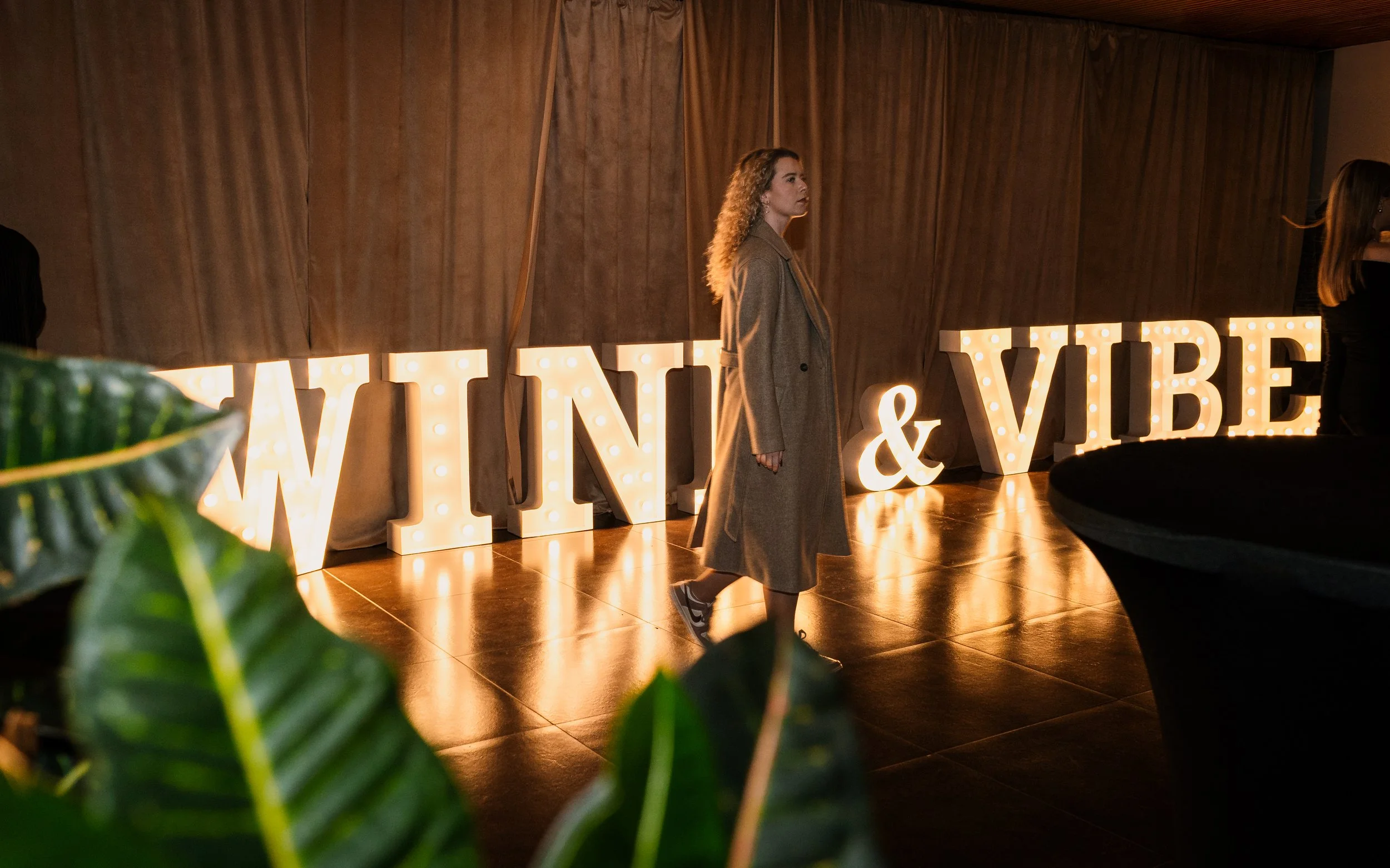 A woman with curly hair wearing a beige coat walking past illuminated decorative letters that spell out 'WINE & VIBE' at an indoor event, with dark curtains in the background and a green plant in the foreground.