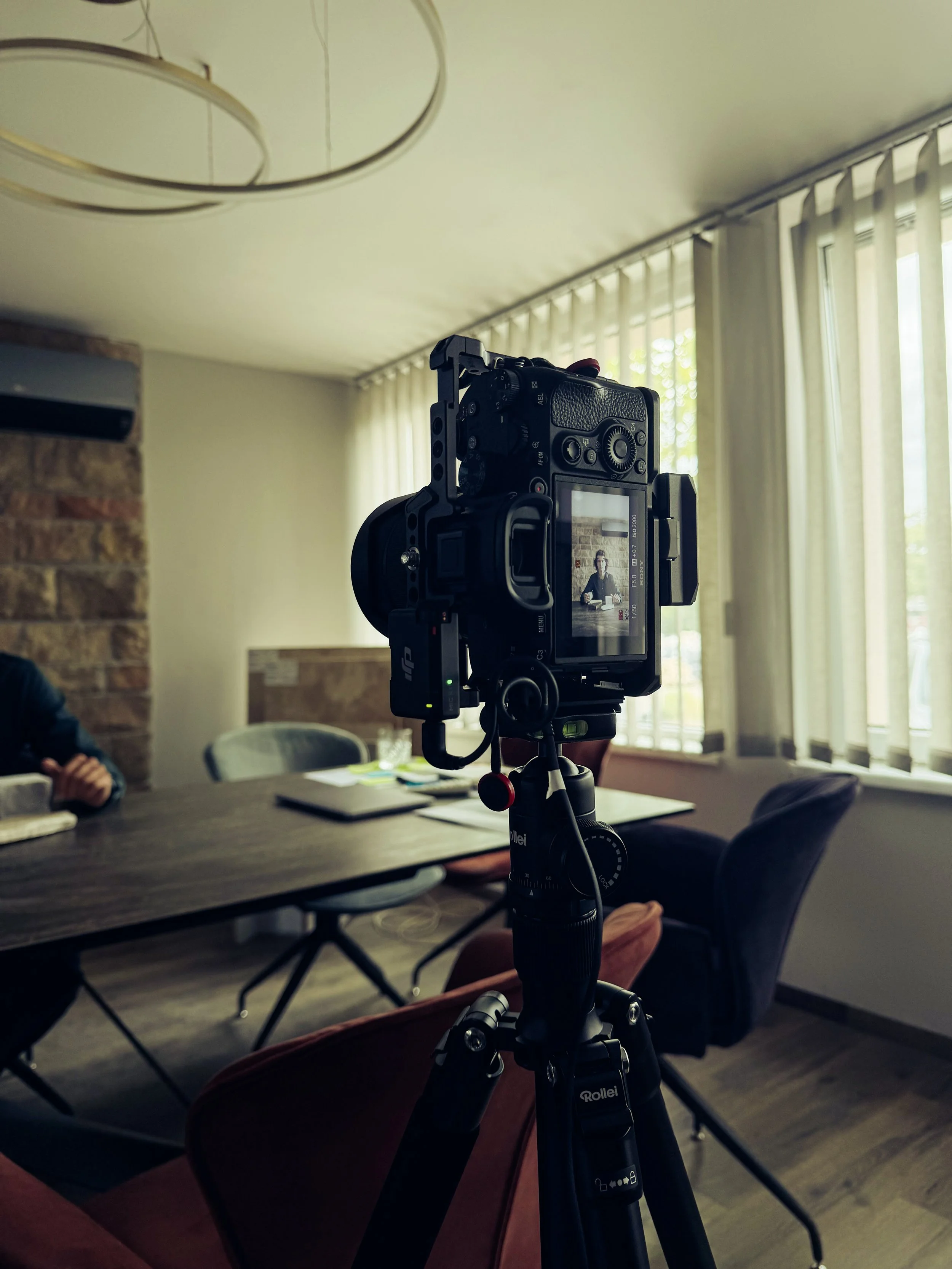 Indoor conference room with a camera mounted on a tripod aimed at a table with people sitting around it. The room has large windows with vertical blinds and an exposed brick wall.