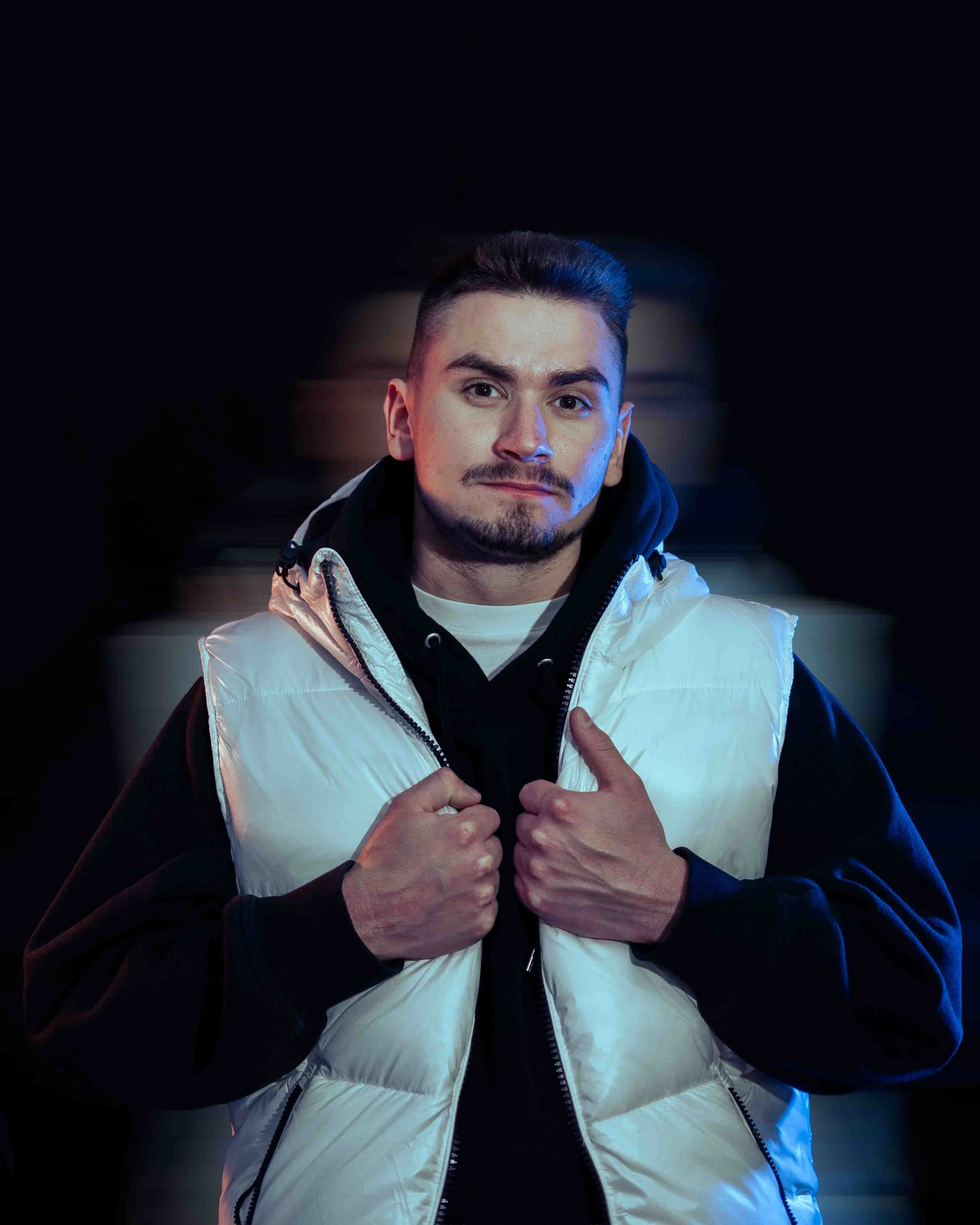 A young man with short dark hair, facial hair, and light skin, wearing a white jacket with black accents and a black hoodie underneath. He is holding the zipper of his jacket with both hands and smiling slightly. The background is dark with a blue tint.