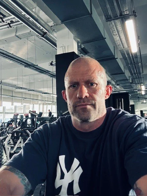 Samuel Pontin a dark t-shirt at the gym, with exercise equipment and large windows in the background.