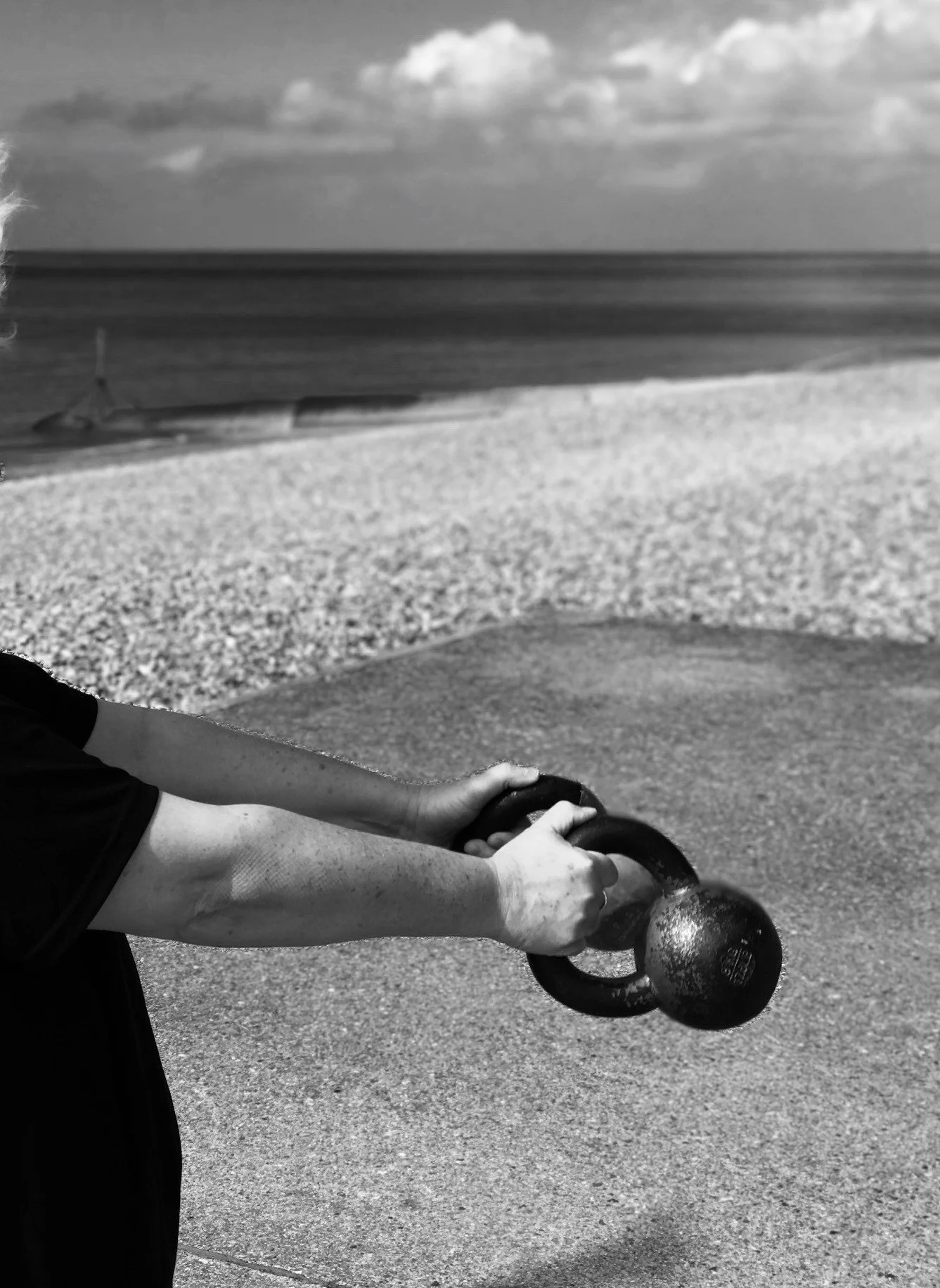 Person holding a kettlebell on a paved surface near a beach, with the ocean and clouds in the background, black and white photo.