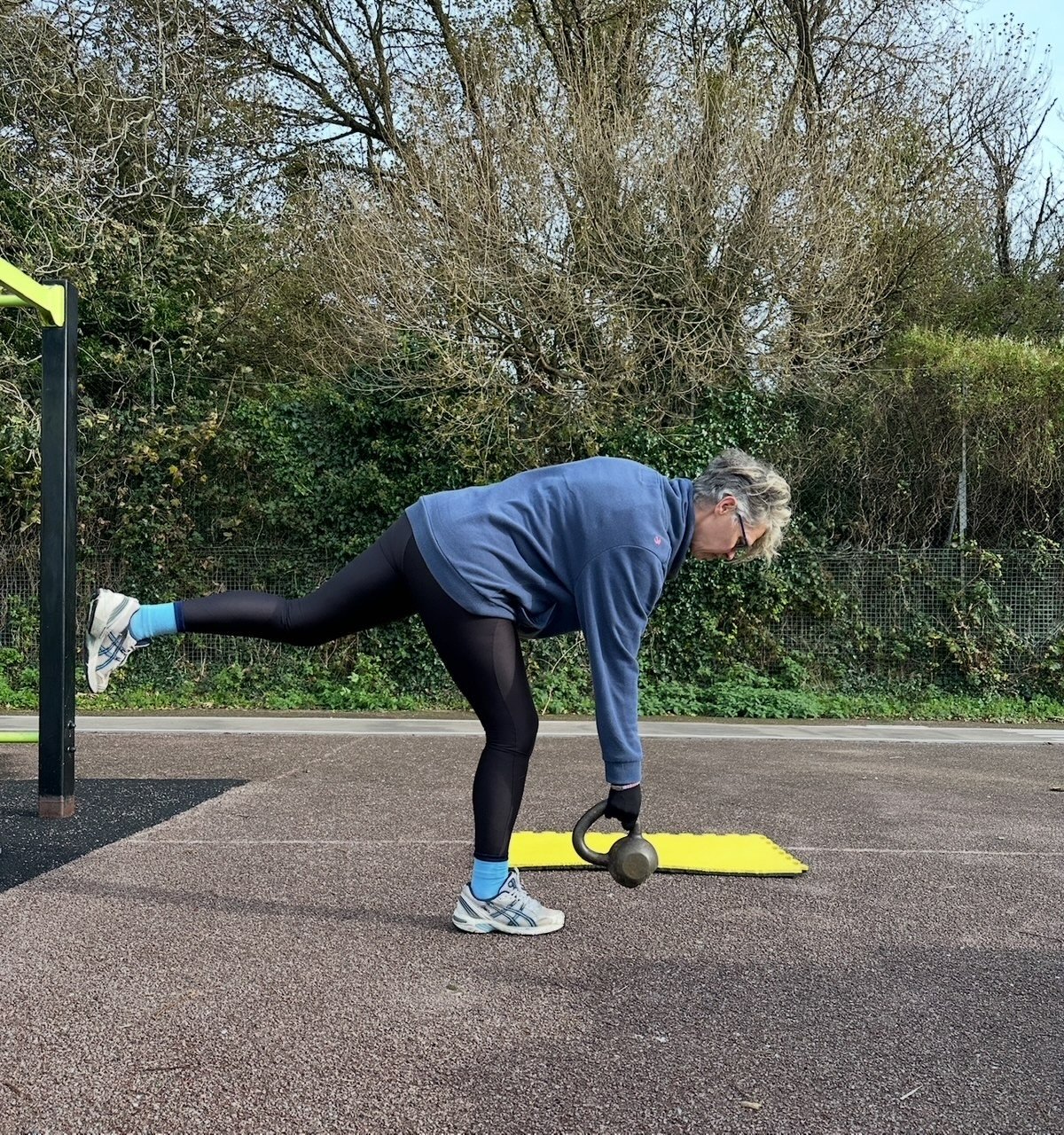 A woman exercising outdoors on a paved surface, balancing on her right leg with her left leg extended behind her, holding a kettlebell in her right hand, with a yellow exercise mat nearby, trees and bushes in the background.