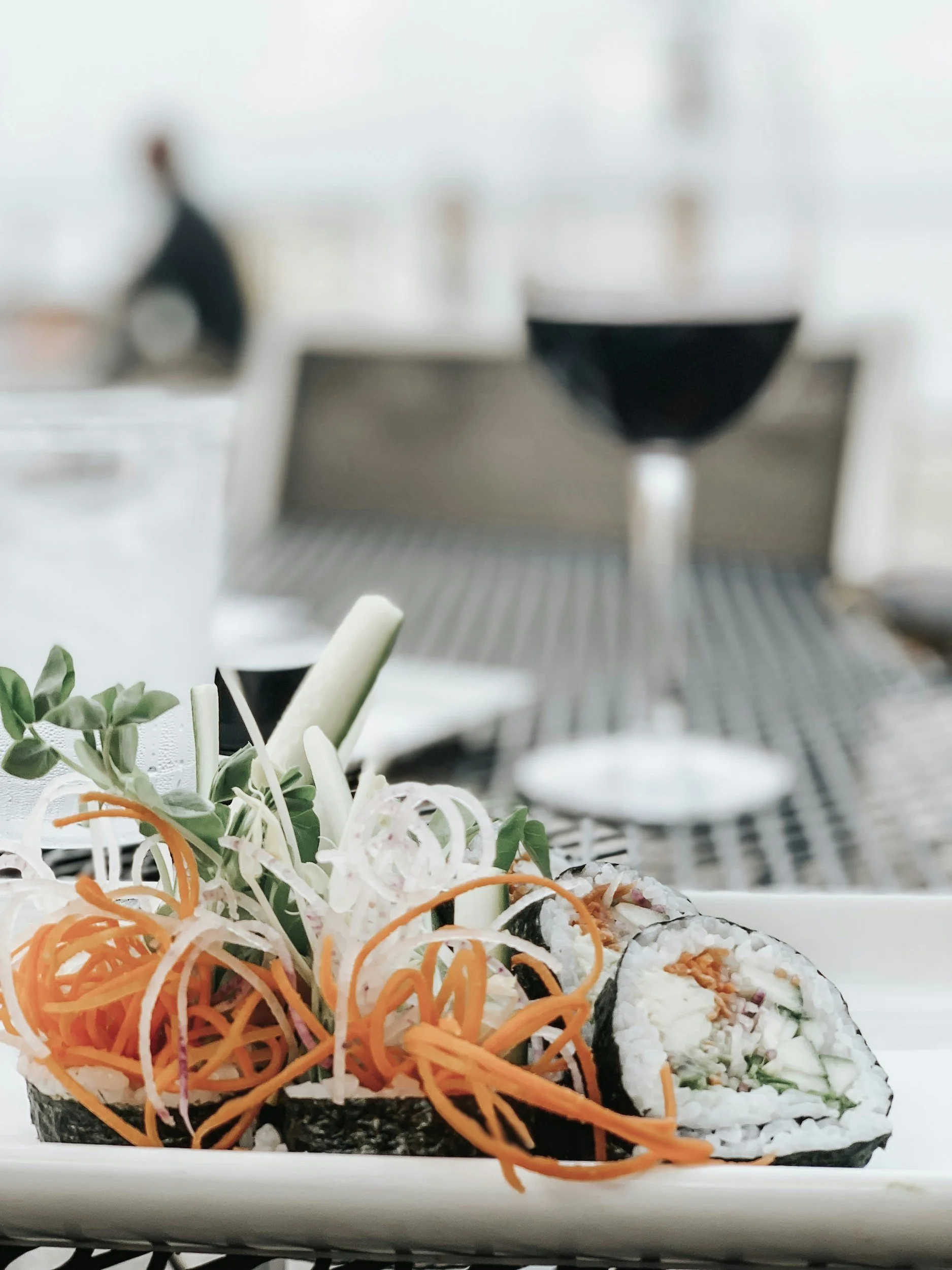 Plate of sushi rolls with shredded carrots, cucumber, and garnishes with a glass of red wine in the background.