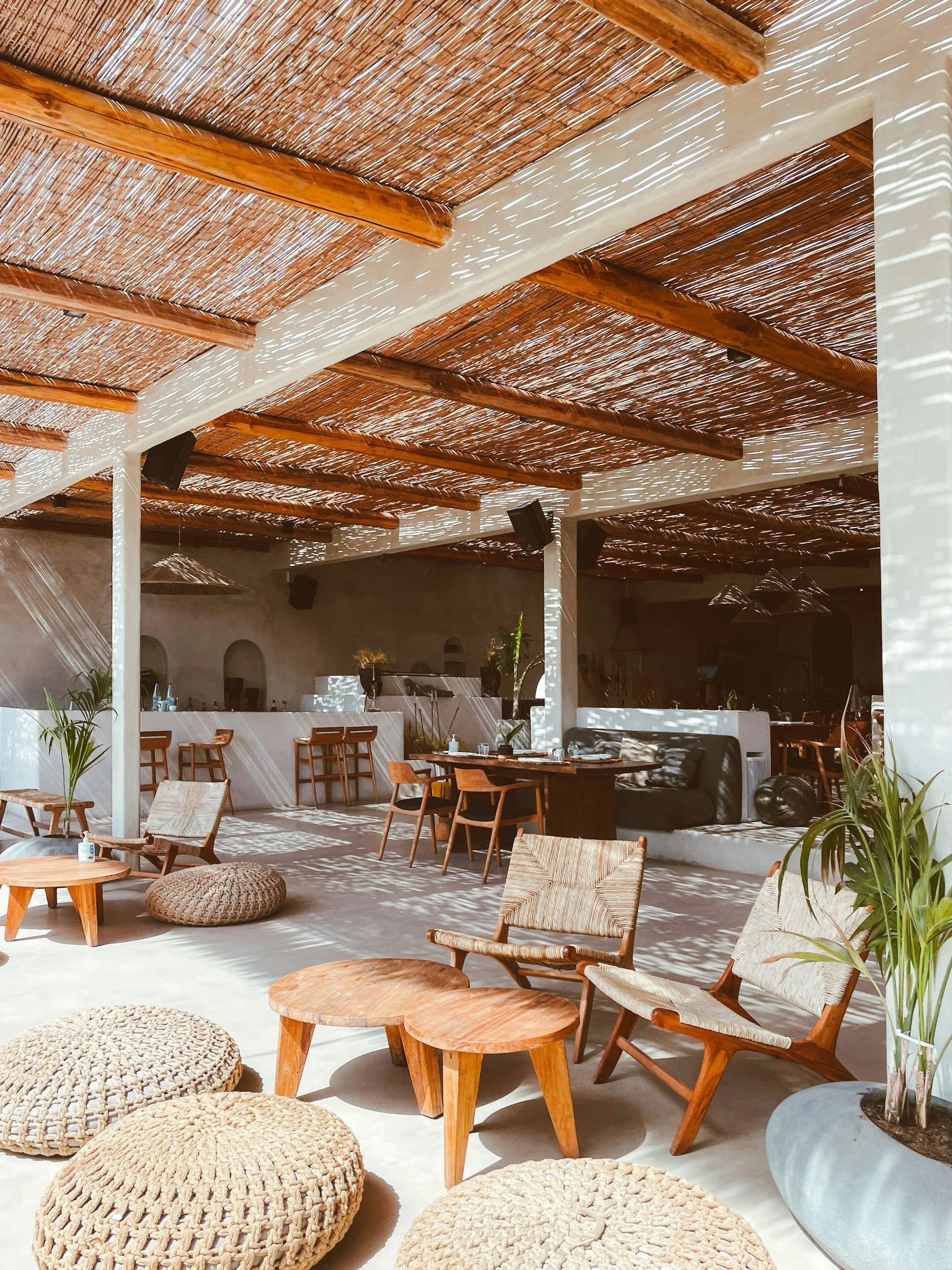 Bright, airy indoor space with wooden furniture, woven rugs, and plants, featuring a thatched ceiling with sunlight casting shadows.