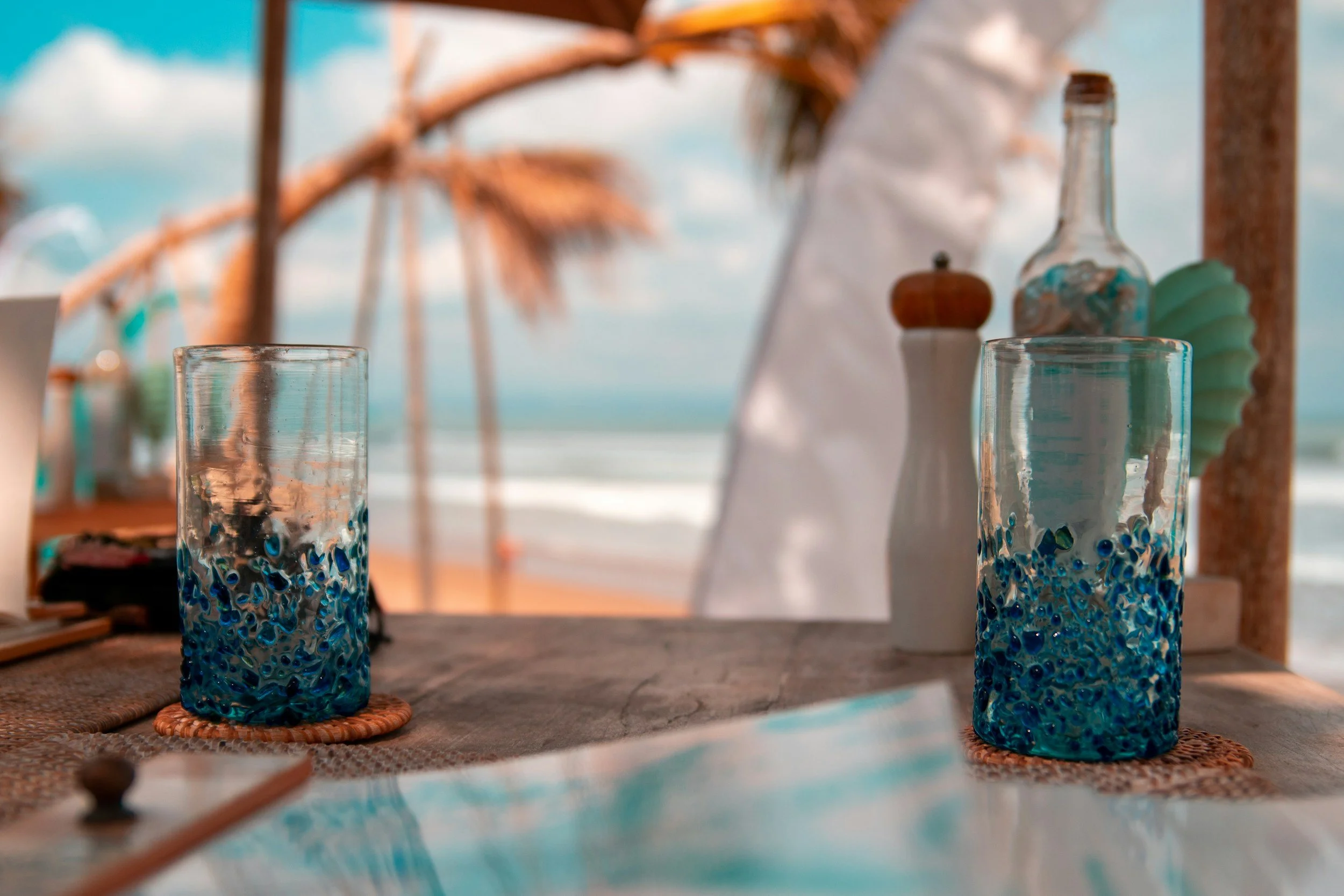 Two tall glasses with blue marble-like patterns filled with water, placed on woven coasters on a wooden table at a beachside restaurant with beach umbrellas and ocean in the background.