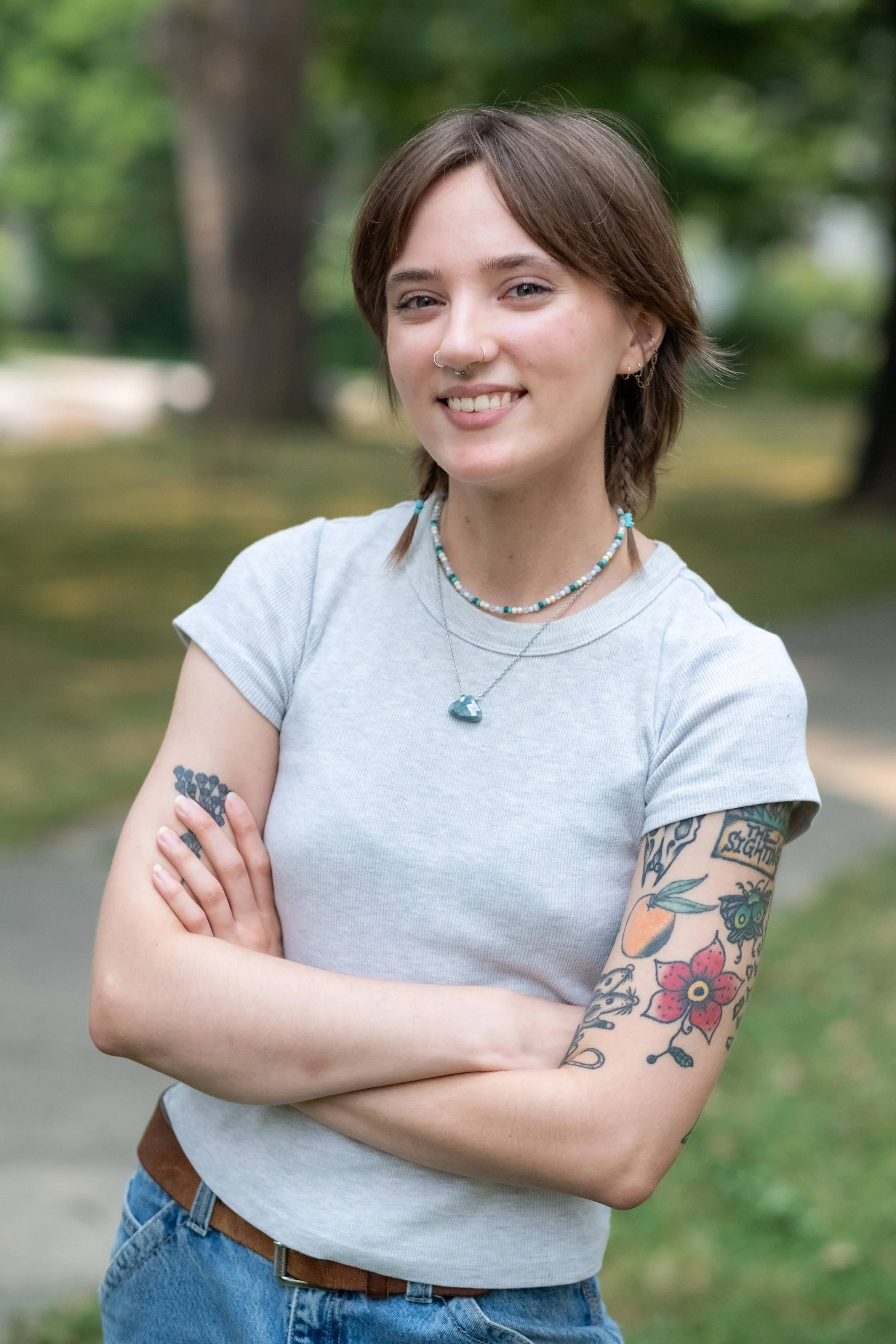A young woman with short hair, wearing a light gray t-shirt, jewelry, and blue jeans, stands with her arms crossed outdoors in a park with green trees in the background.