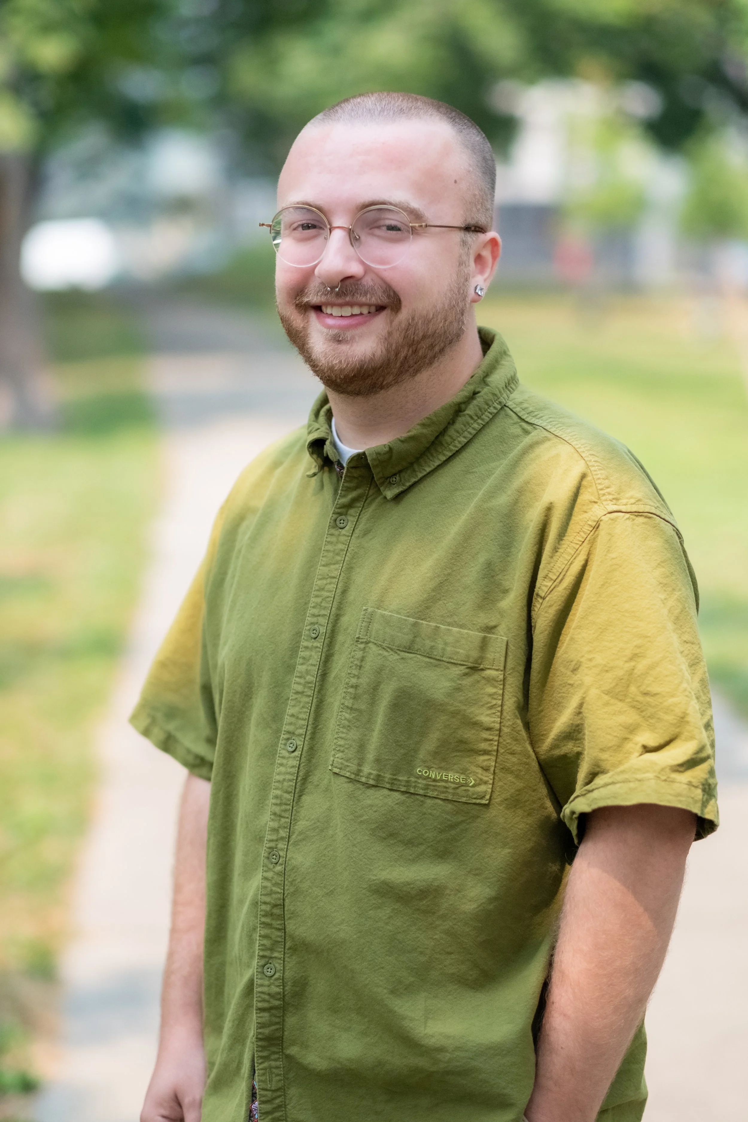 A smiling man with a beard, glasses, and earrings standing outdoors on a path with trees in the background, wearing a green button-up shirt.