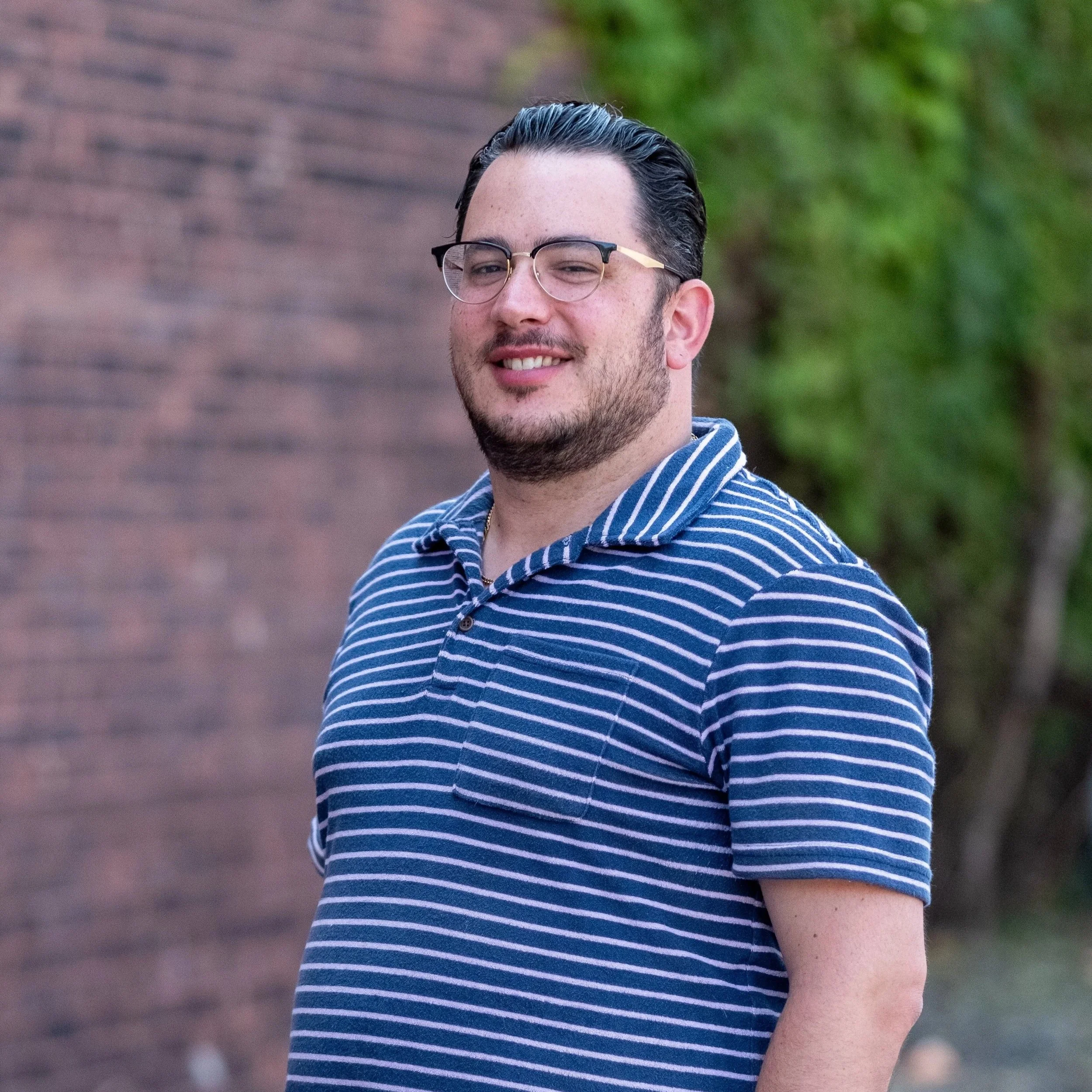 A young man with dark hair, glasses, and a beard, wearing a blue and white striped shirt, standing outdoors with a brick wall and greenery in the background.