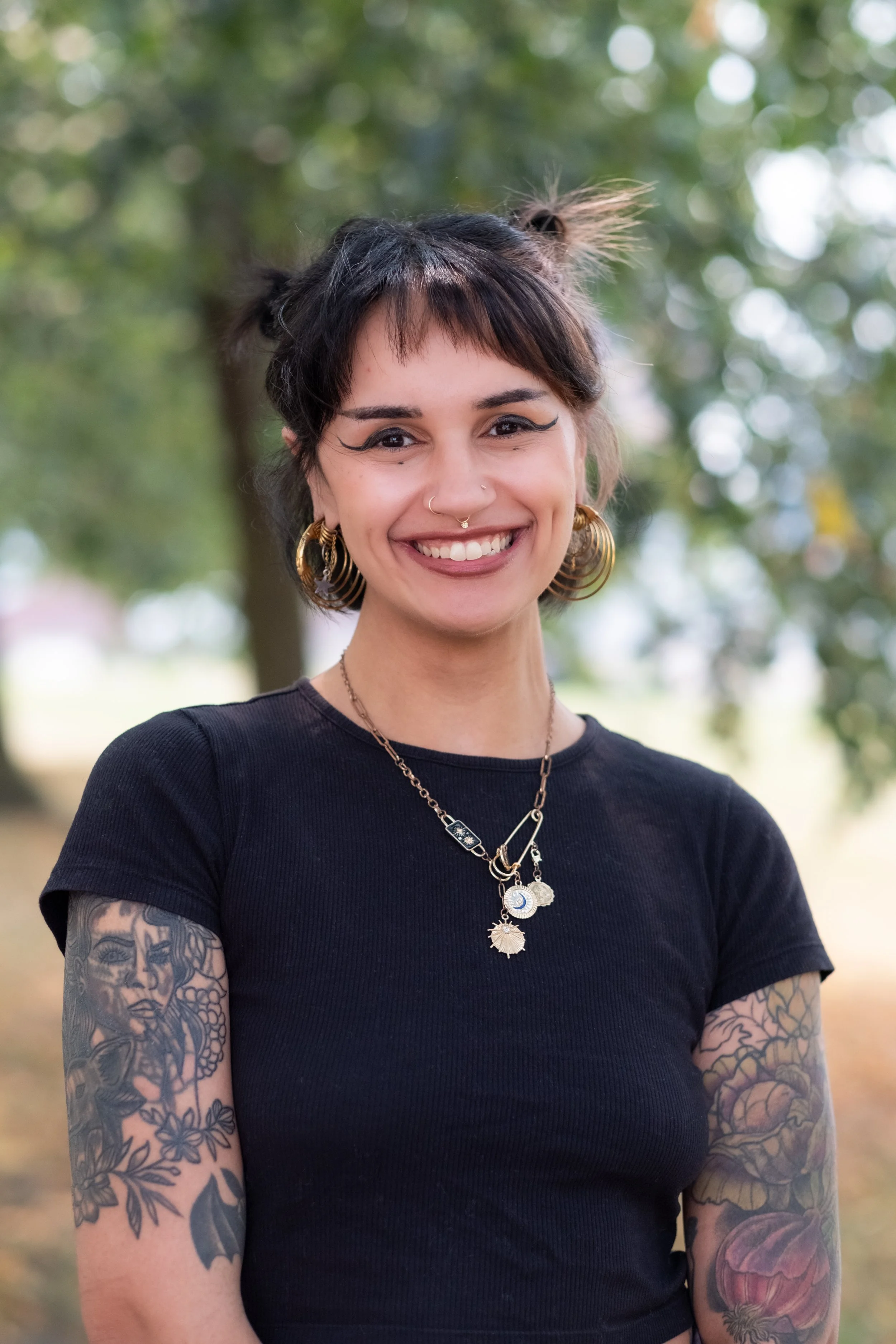 A young woman with tattoos on her arms, wearing a black t-shirt, gold hoop earrings, and layered necklaces, smiling outdoors with an out-of-focus green background.