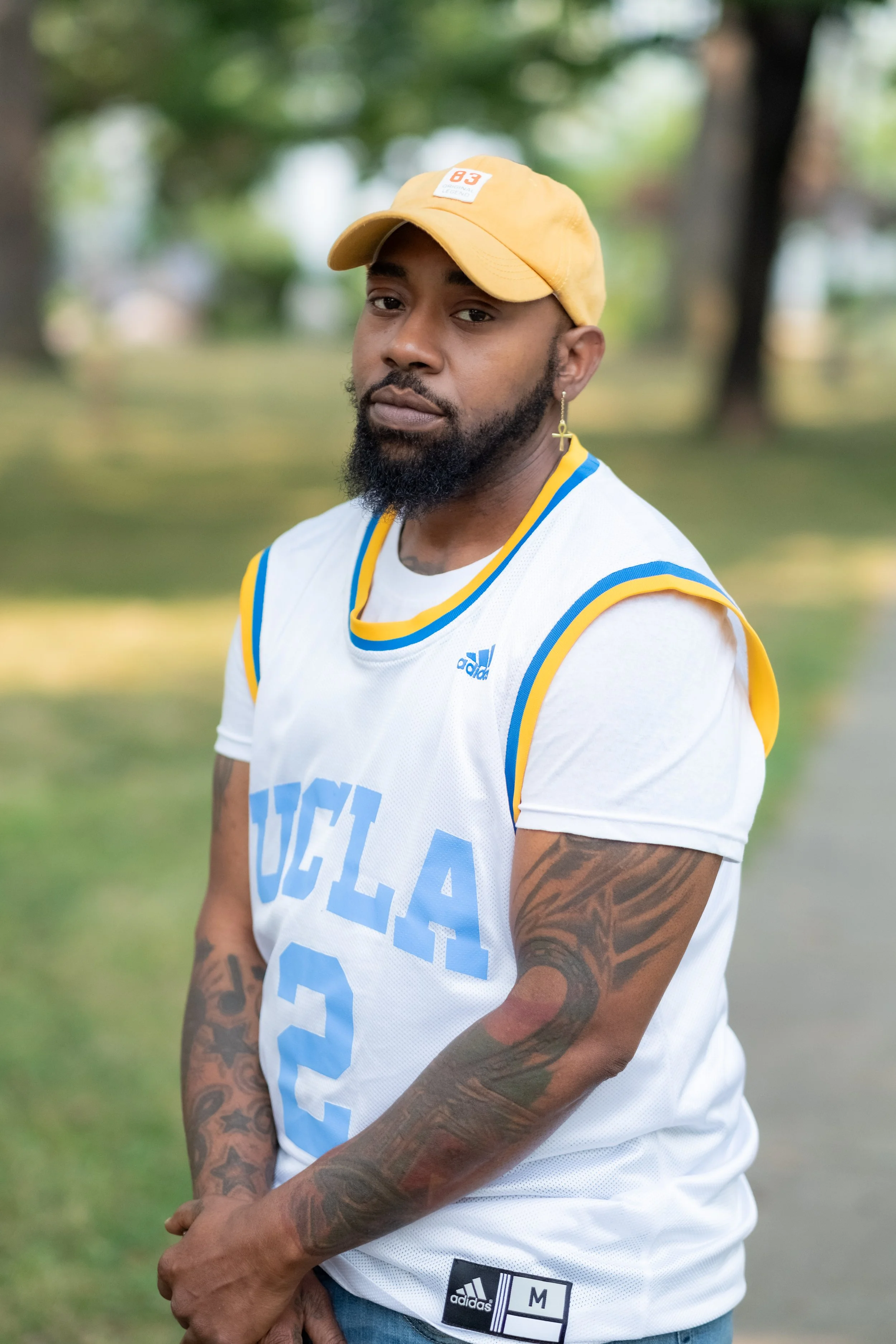 A man wears a yellow baseball cap, a white UCLA basketball jersey, and has tattoos on his arms. He has a beard, an earring, and is outdoors with trees in the background.