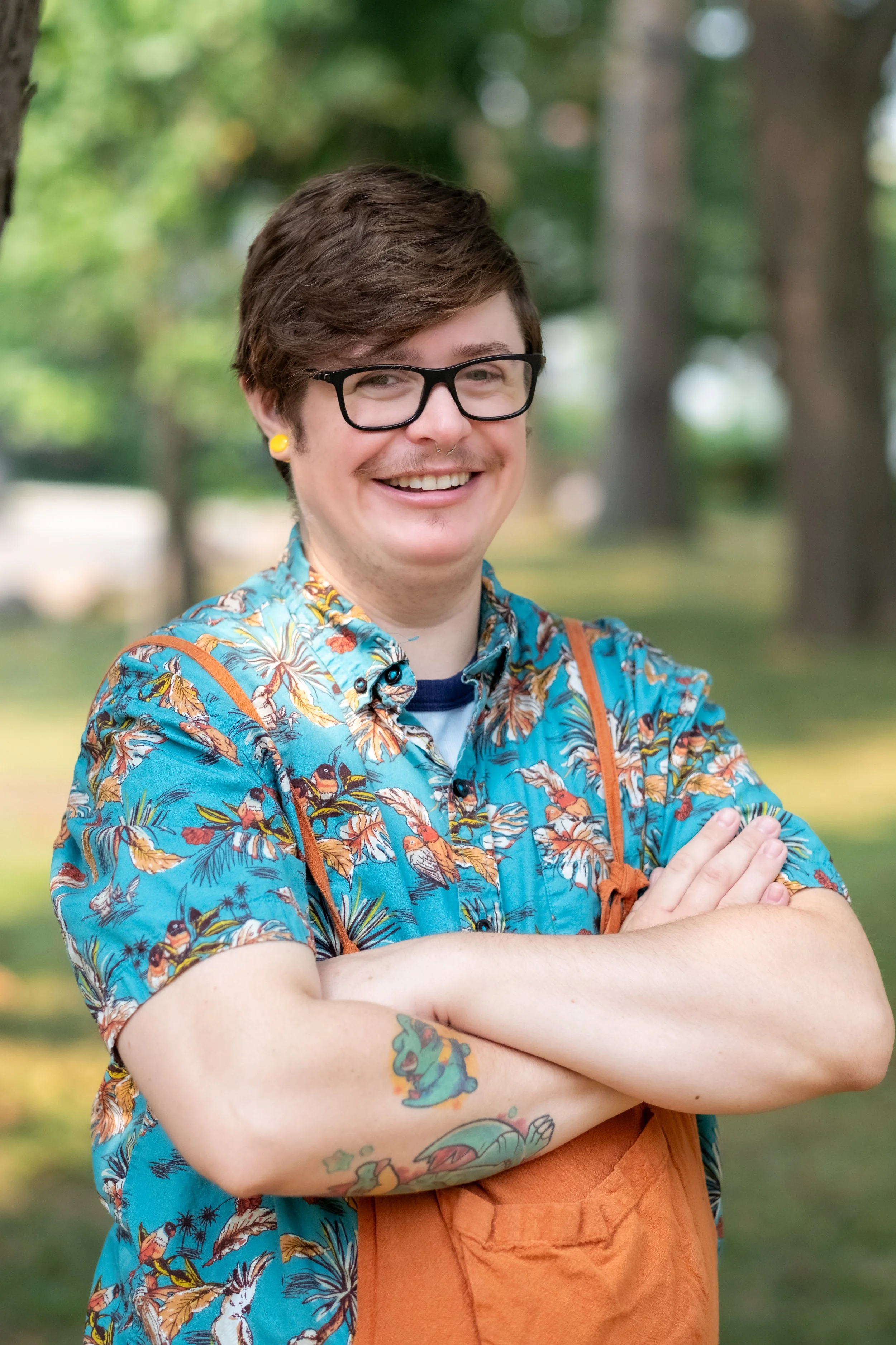 A smiling young man with short brown hair, black glasses, and a nose ring, wearing a blue Hawaiian shirt with a tropical bird pattern, tattoos on his arms, and orange overalls, standing outdoors in a park.