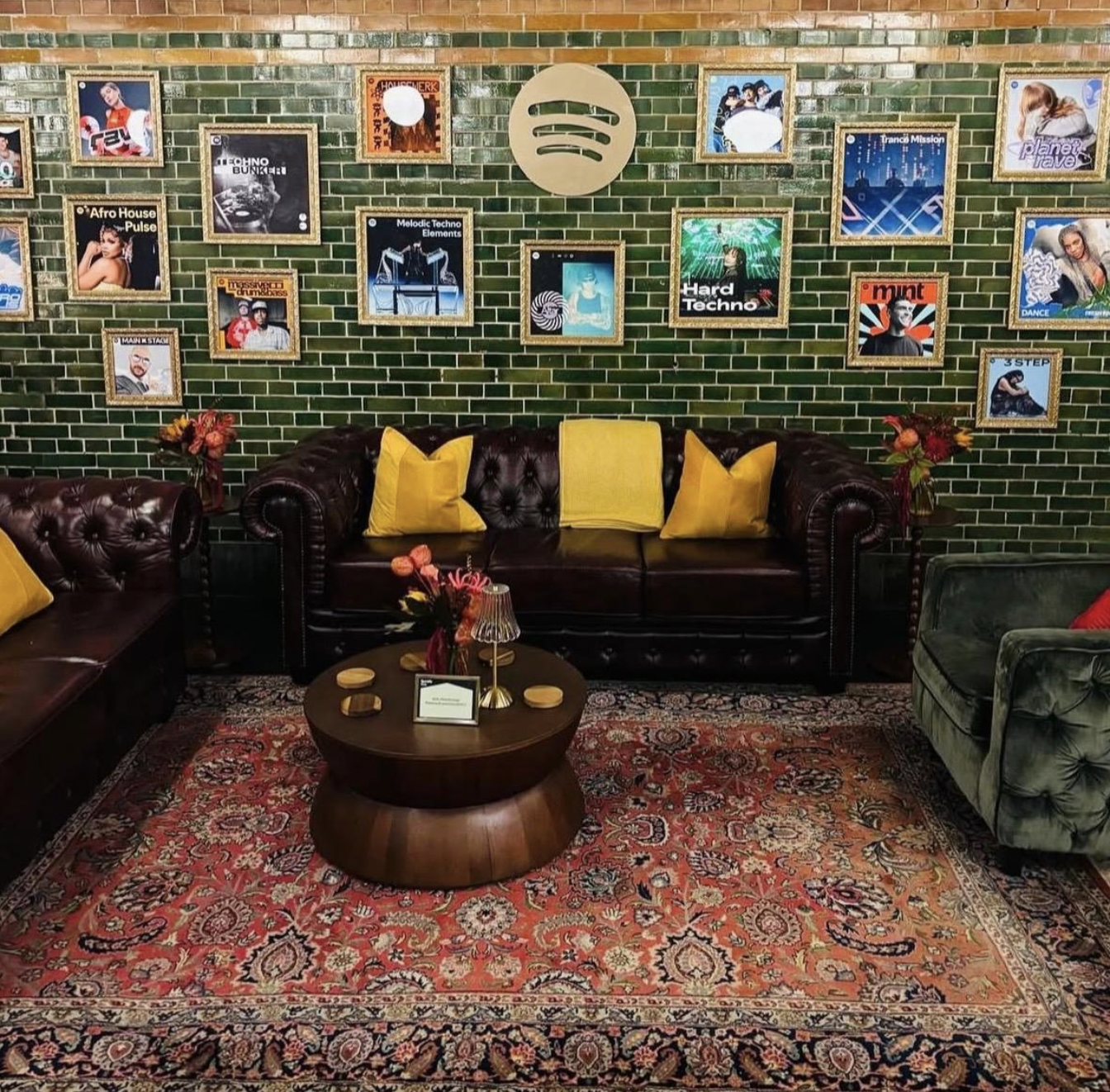 A cozy living room with a green brick wall decorated with framed posters of music genres and artists, a dark brown leather sofa with yellow pillows, a round wooden coffee table with small decorative coasters and a lamp, a patterned area rug, and small side tables with flower arrangements.