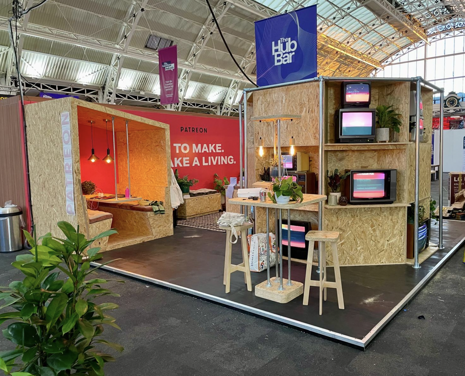 A booth at a convention or trade show features a wooden structure with multiple vintage televisions, a small table, and plants. The booth has signs for 'The Hub Bar' and 'Patreon'  Live show producer, set designer, table scaping, event producer, even