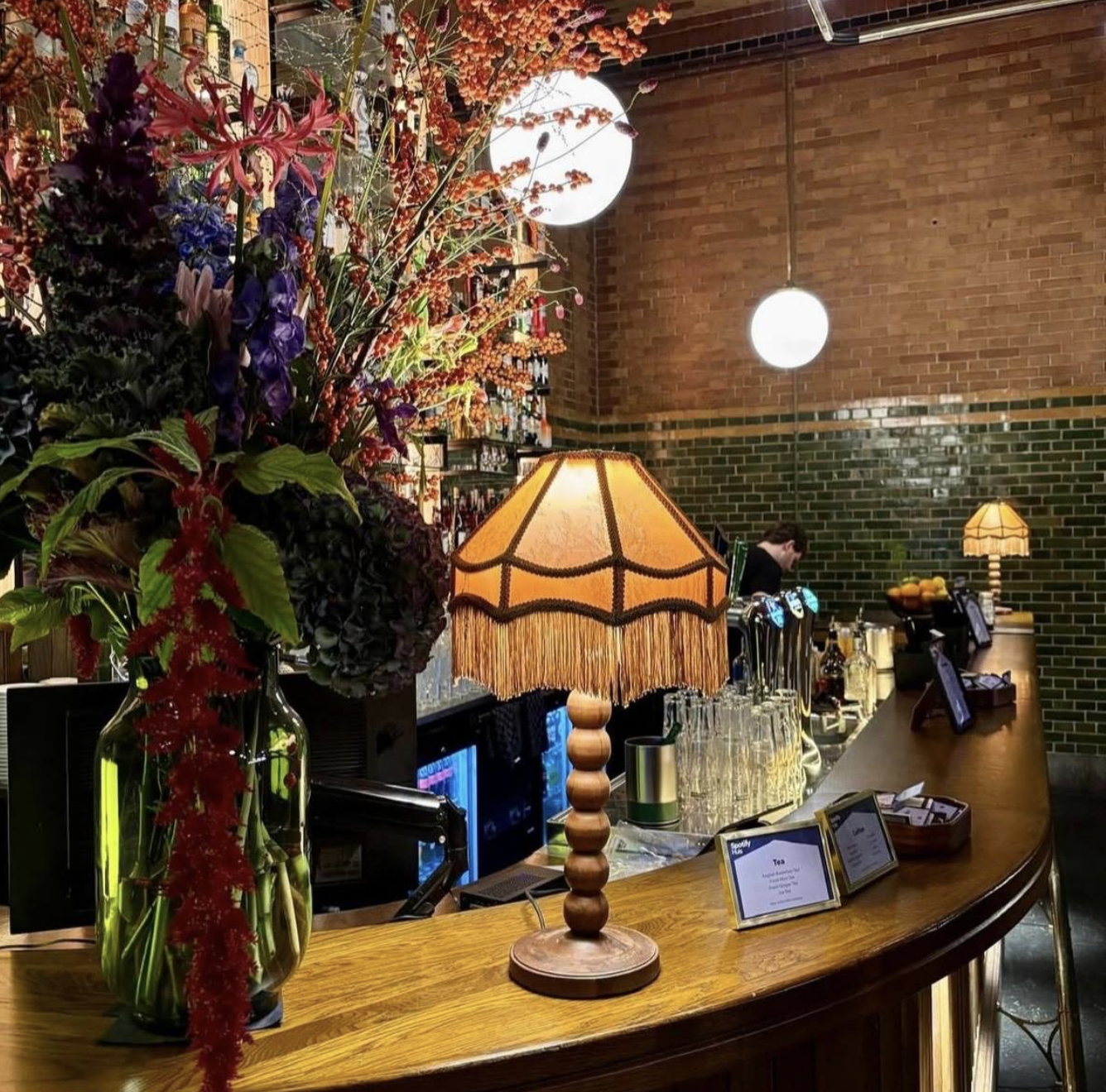 A bar with a large colorful flower arrangement, two vintage-style table lamps, and a brick wall background. Live show producer, set designer, table scaping, event producer, event consultant, freelance, private, corp, london, worldwide, europe Spotiy