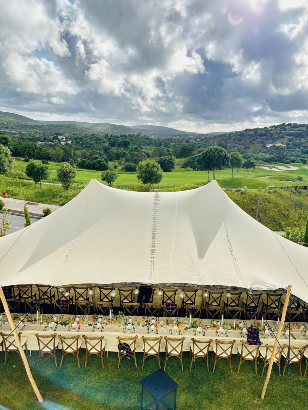 Long outdoor table set for a banquet under a large white canopy on a grassy area. The table is decorated with flowers and filled with place settings, and surrounded by chairs. In the background, there are green rolling hills, trees, and a partly cloudy sky.