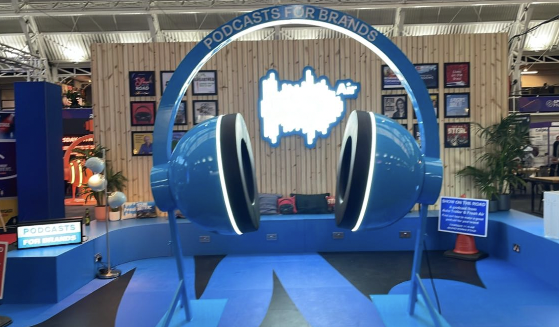 A blue and white podcast-themed exhibit with large headphones and a city skyline logo, featuring signs that say 'Podcasts for Brands' and a seating area with pillows, in a convention or expo setting.