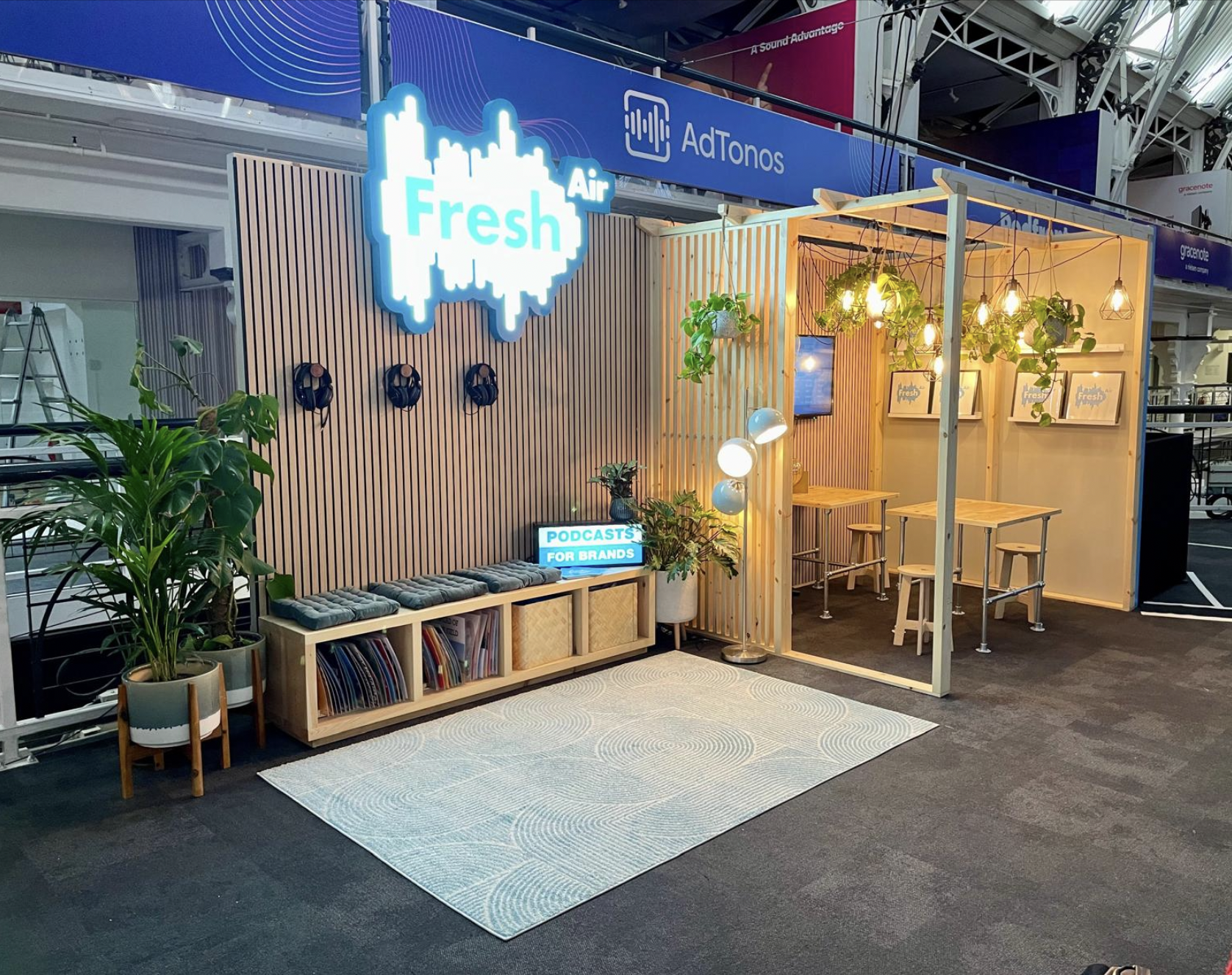 Exhibit booth with a large illuminated sign that says 'Fresh' Air and a smaller screen saying 'Podcasts for Brands', decorated with plants, hanging lights, and framed prints. Live show producer, set designer, table scaping, event producer, event con