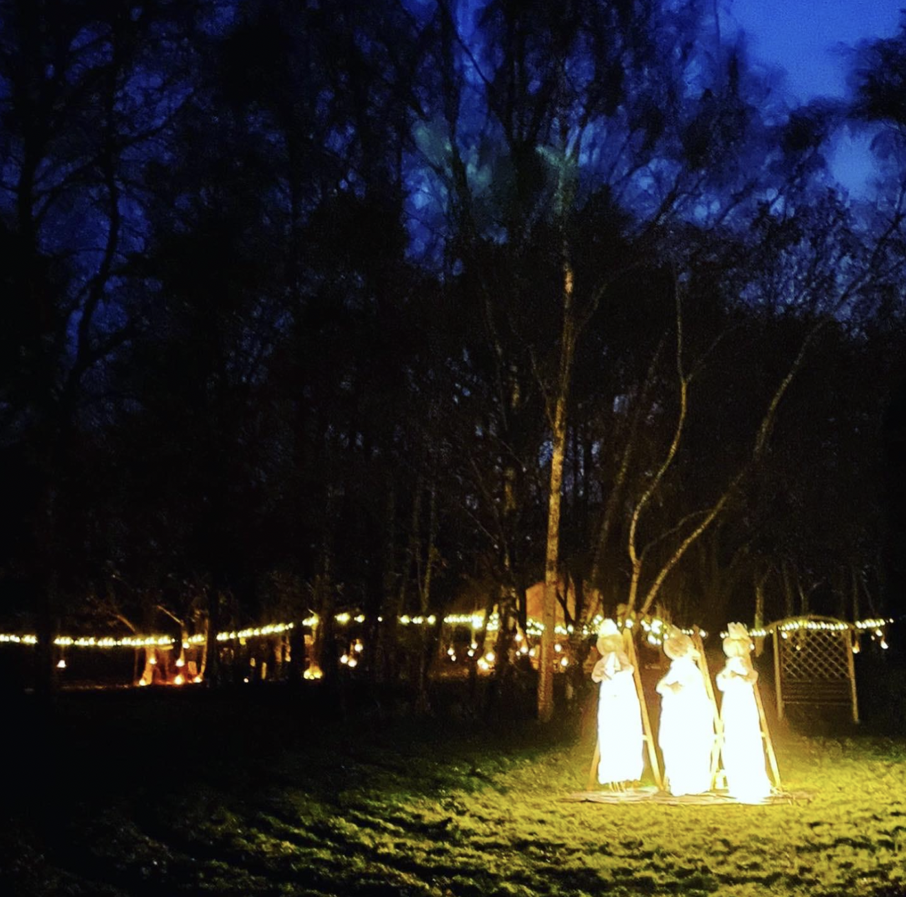 Nighttime outdoor scene with three illuminated angel statues on a grassy area, trees in the background with string lights, and a wooden fence with a gate. Live show producer, set designer, table scaping, event producer, event consultant, freelance, p
