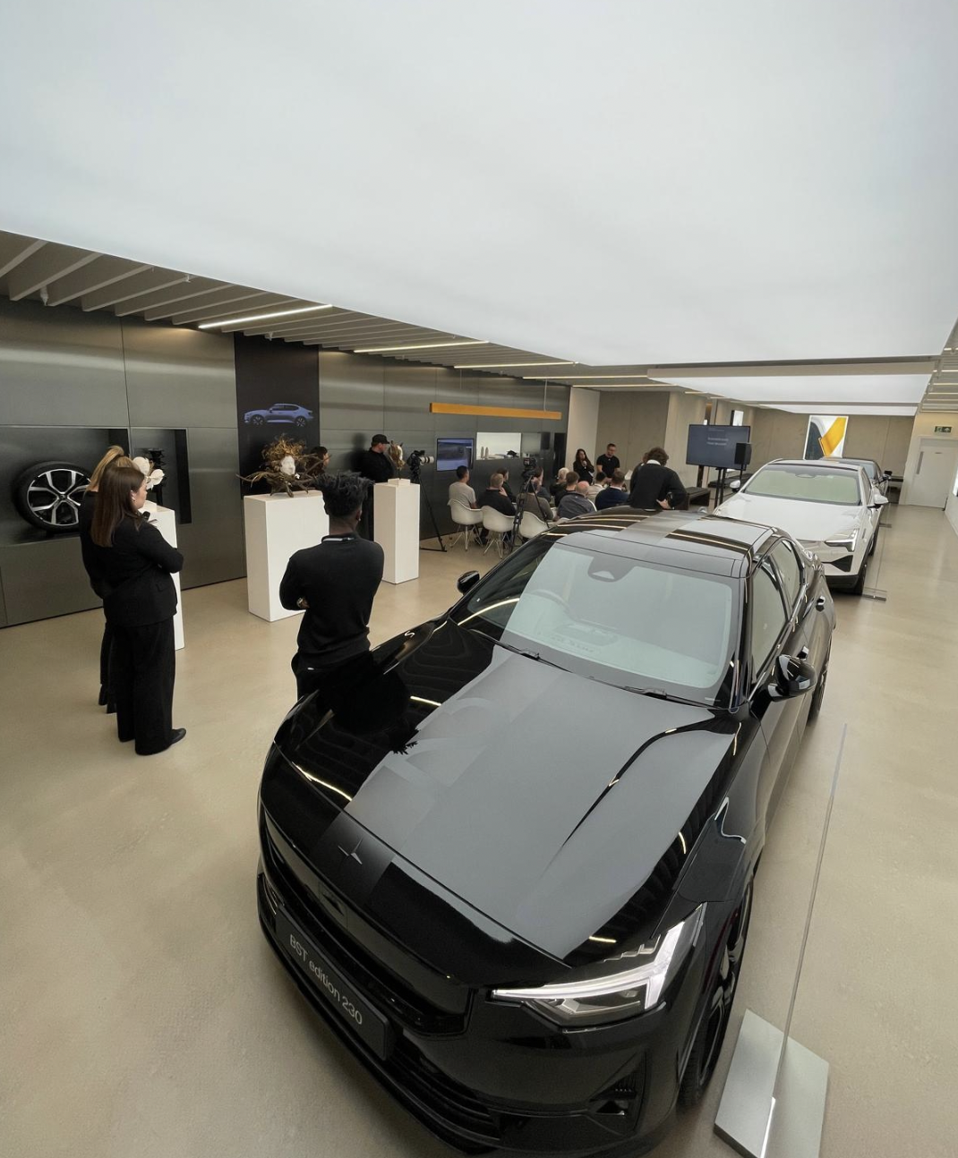 A showroom filled with new cars and people attending a presentation or event, with a black electric car labeled 'Best Edition 230' in the foreground.