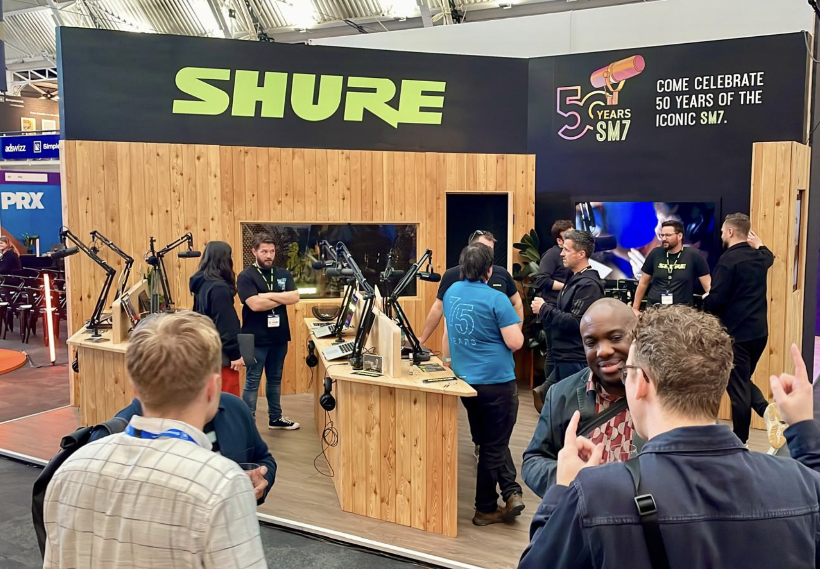 A booth at an event celebrating the 50th anniversary of the Shure SM7 microphone, with multiple people engaging and discussing around wooden display tables equipped with computers and microphones. Live show producer, set designer, table scaping, even