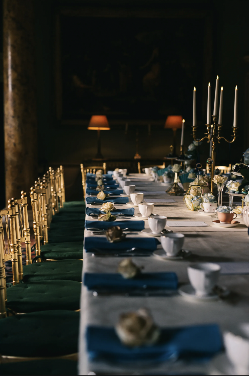 Elegant dining table set for a formal event with white tablecloth, china teacups, blue napkins, and tall candelabras with white candles, in a dimly lit room with paintings and warm lighting.