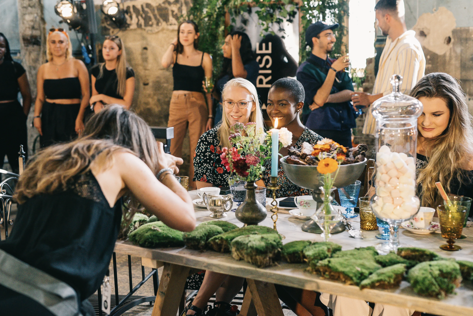 Group of women celebrating a birthday with a decorated cake, surrounded by snacks and drinks, in a rustic indoor setting.