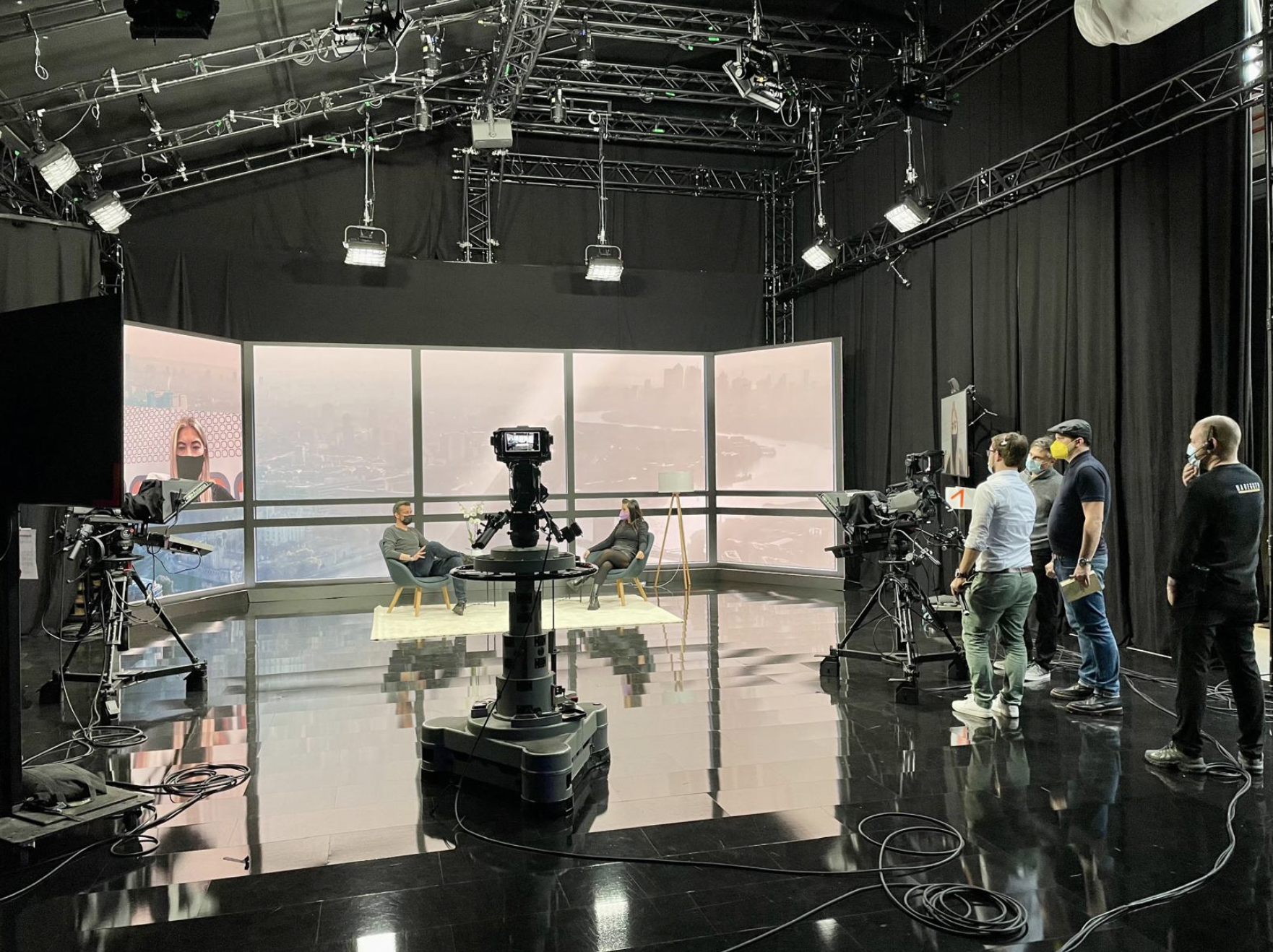 Television studio with a curved large screen displaying a cityscape, two people seated and talking, several crew members standing nearby, professional cameras and lighting equipment present, black curtains and ceiling film producer, event producer,