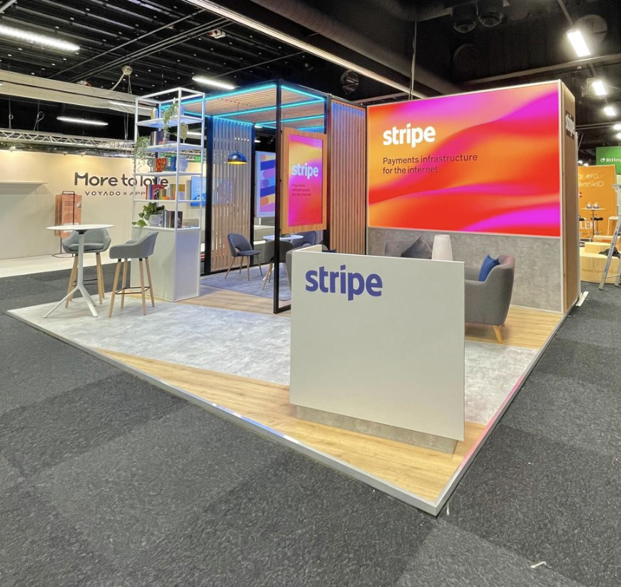 Trade show booth for Stripe featuring a large digital screen with orange and pink graphics, a white table with chairs, a gray sofa, and decorative shelving with plants. Live show producer, set designer, table scaping, event producer, event consultant