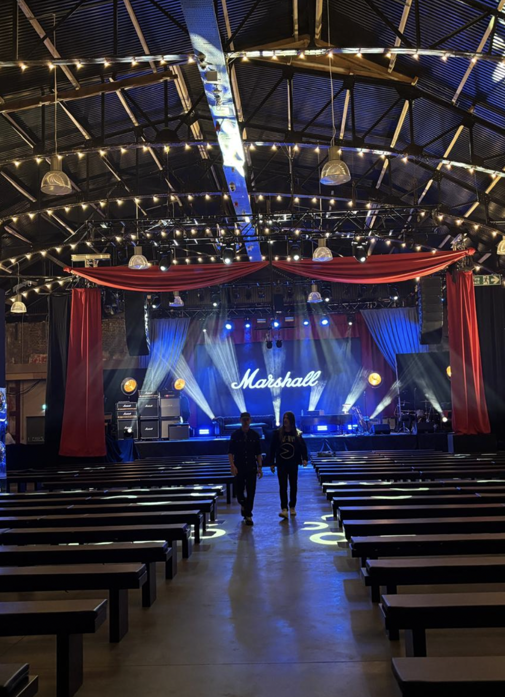Concert venue with a stage set for a performance, illuminated with blue and white lights, featuring the word 'Marshall' on the backdrop, red curtains, and empty benches facing the stage, with two people walking towards the stage.