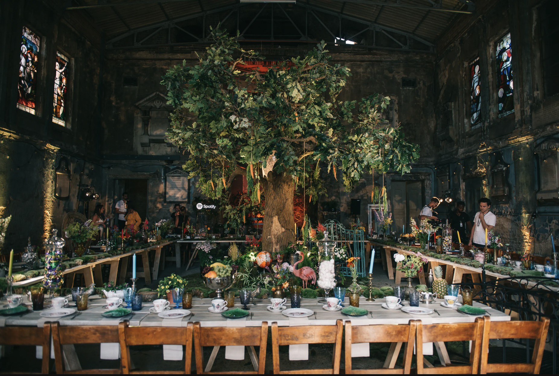Indoor event space with a large central tree and long tables decorated with flowers, glassware, and tableware, set for a celebration or gathering. People are standing and preparing around the tables, with stained-glass windows and a dark, aged interior.
