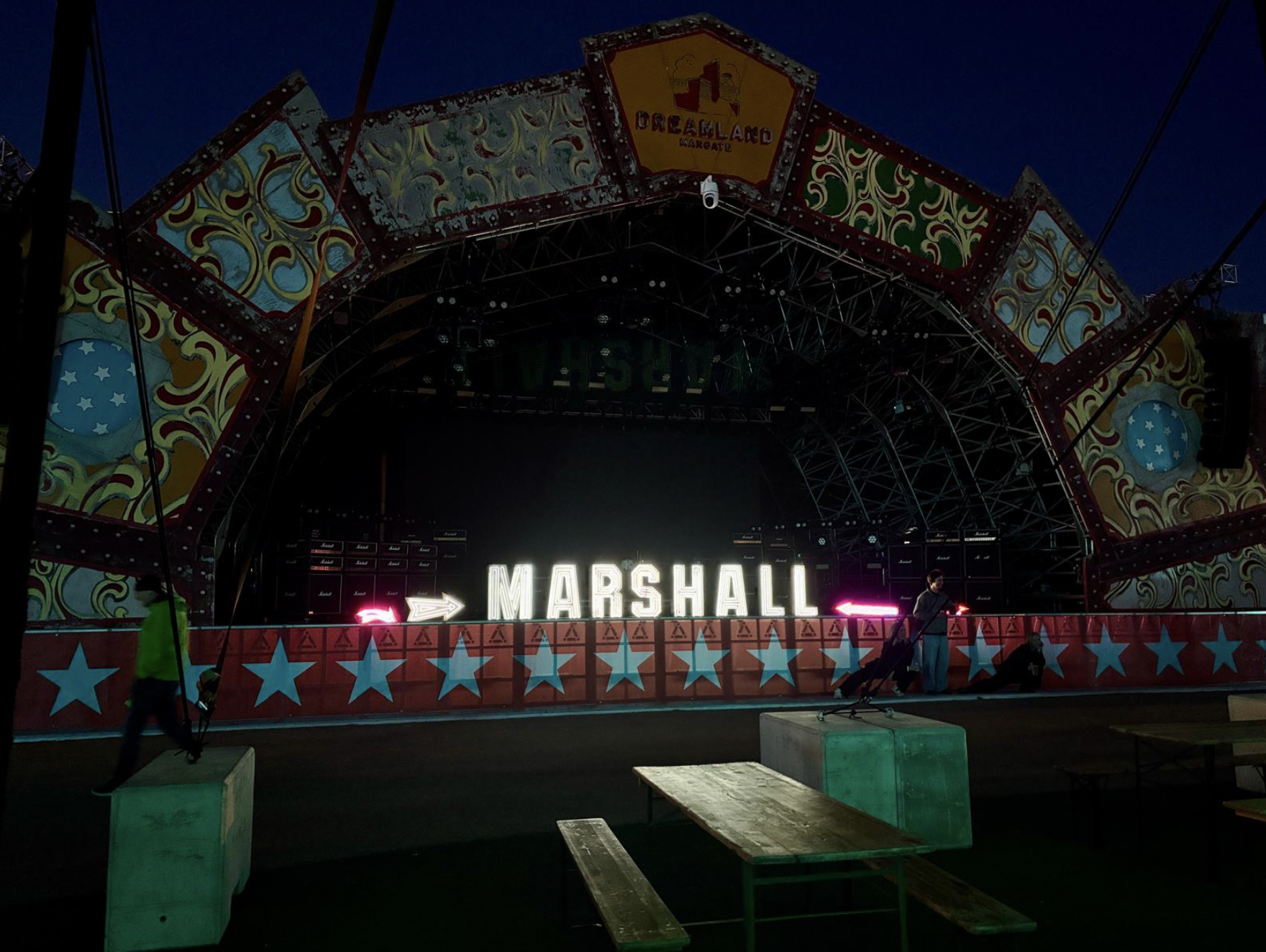 Empty carnival or concert stage with a large illuminated sign that reads 'MARSHALL' and a colorful, decorated arch overhead. The scene is set at dusk or night, with a person and a dog near the stage and some benches in front. Live show producer