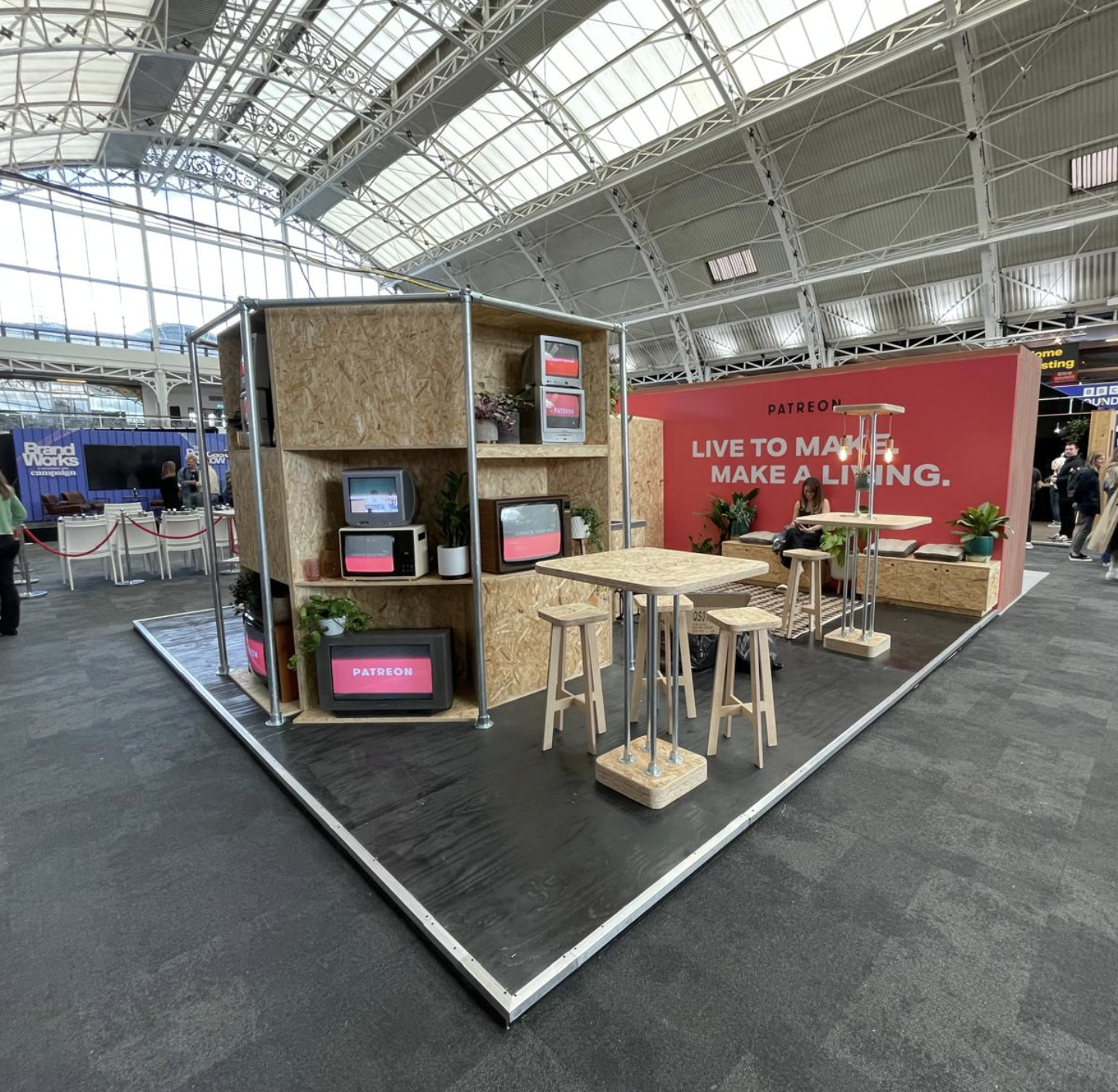 Patreon exhibition booth at a conference with retro TVs, wooden furniture, and a red wall with the slogan "Live to make, make a living."