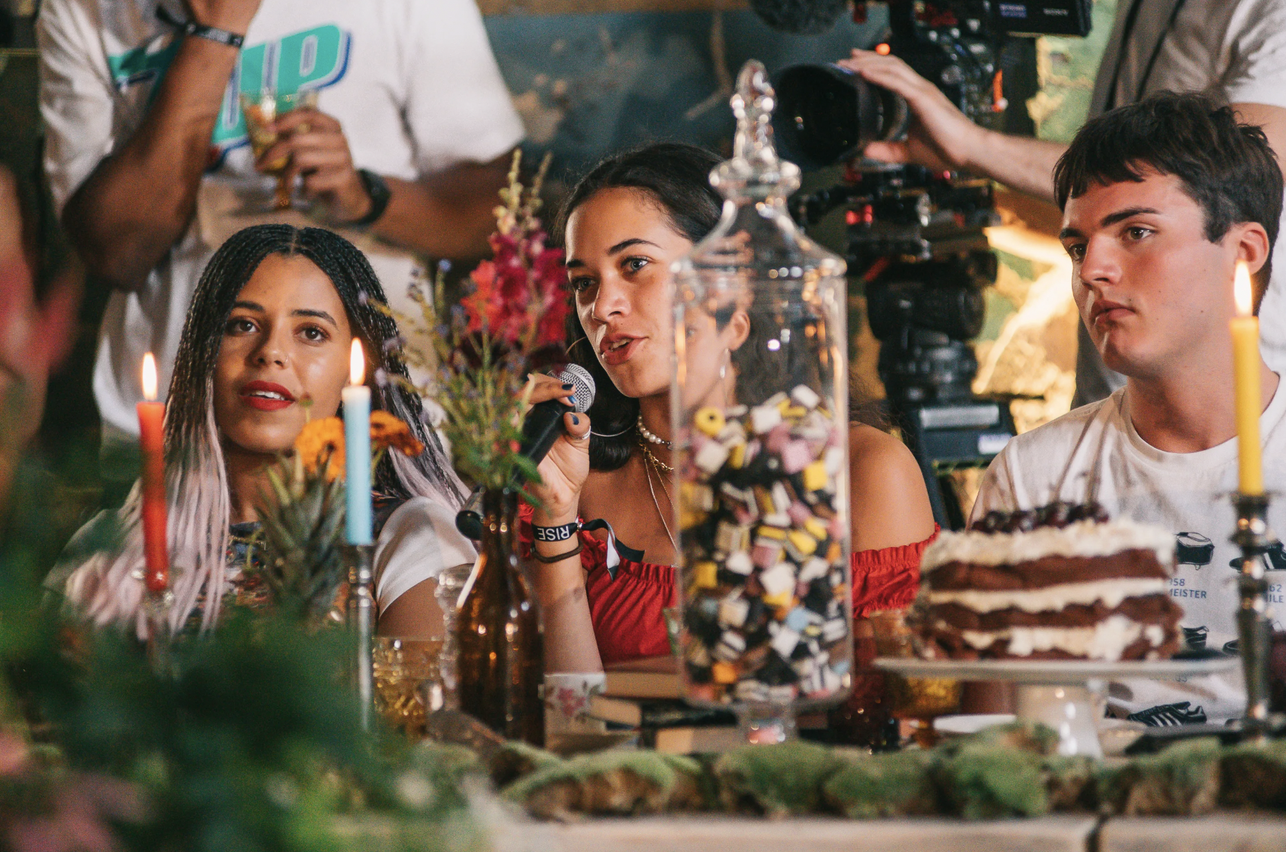 Three young people sitting at a decorated table with candles, flowers, and a cake, surrounded by cameras and crew, including one woman holding a microphone, during a filming or photo shoot.
