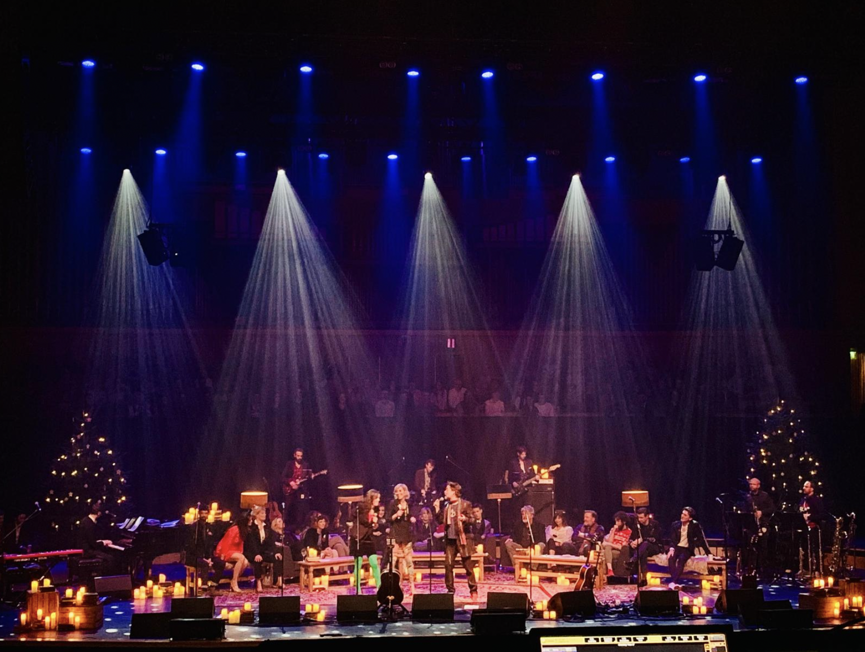 A stage decorated with Christmas trees and candles, featuring a large group of singers and musicians performing under colorful stage lights. Live show producer, set designer, table scaping, event producer, event consultant, rufus wainwright festival