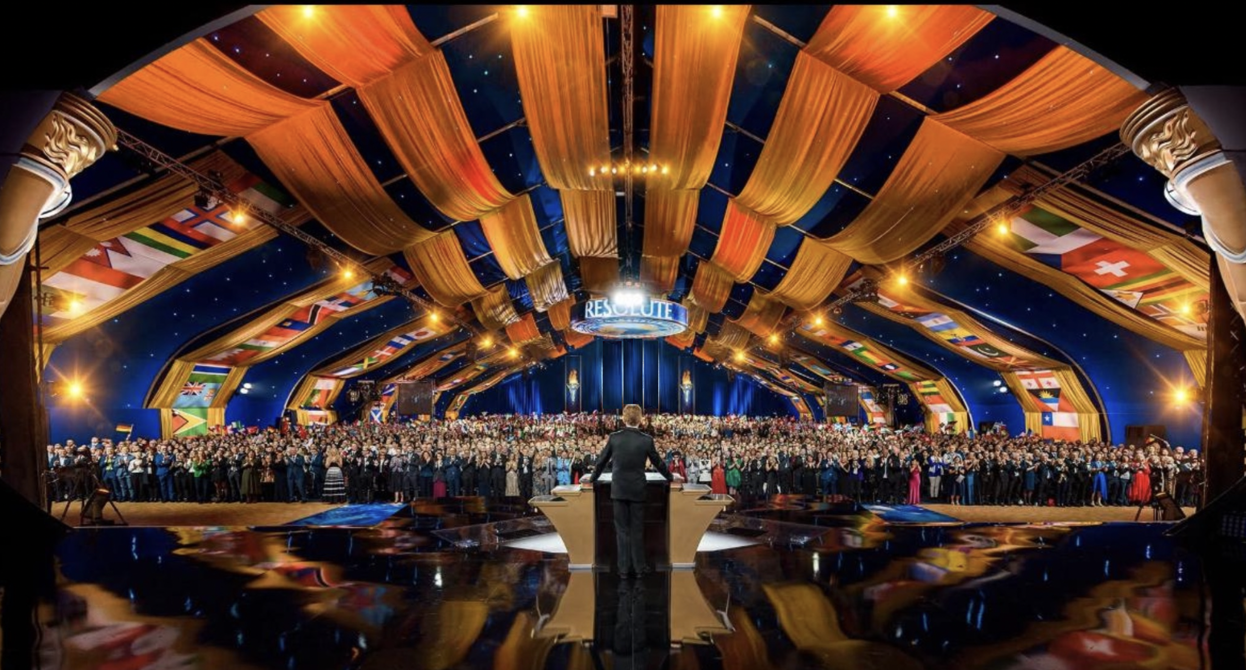Large conference or event with many attendees on stage and a speaker at a podium, under a decorated ceiling with flags from various countries and a sign that reads 'RESOLUTE'.