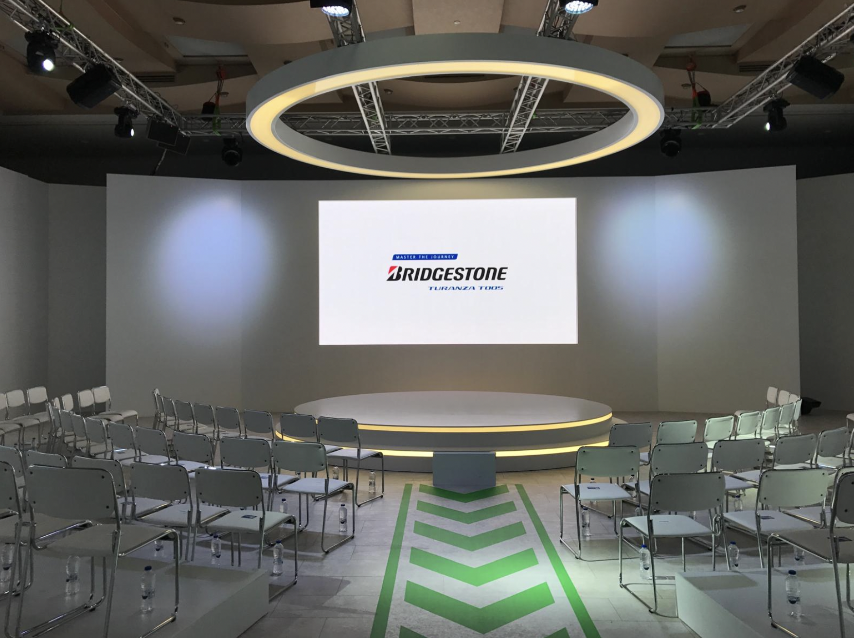An empty conference or presentation room with rows of chairs facing a large screen and a circular stage, illuminated by overhead lights.