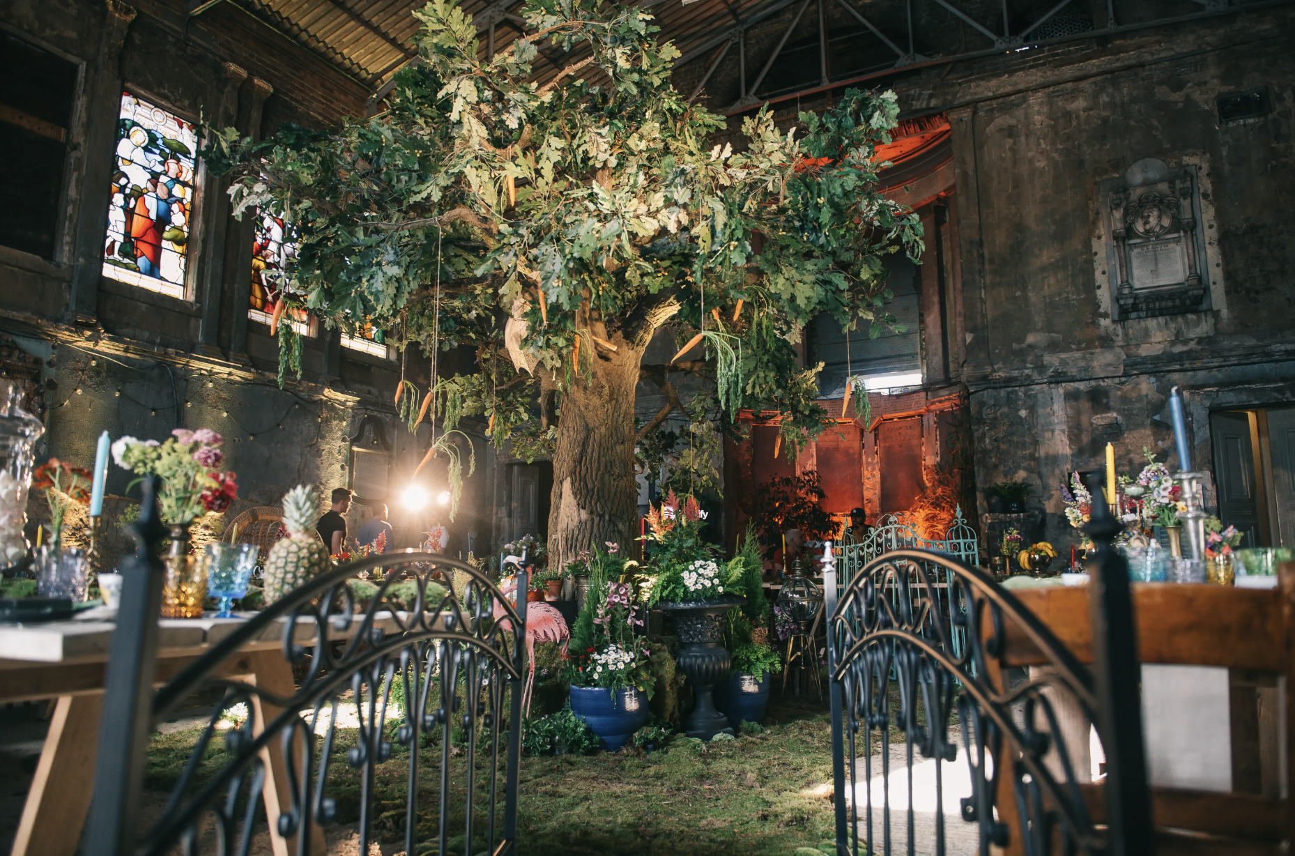Interior of a wedding or event venue decorated with a large indoor tree, flowers, candles, and colorful glassware, with stained glass windows and a rustic stone wall in the background. Live show producer, set designer, table scaping, event producer,