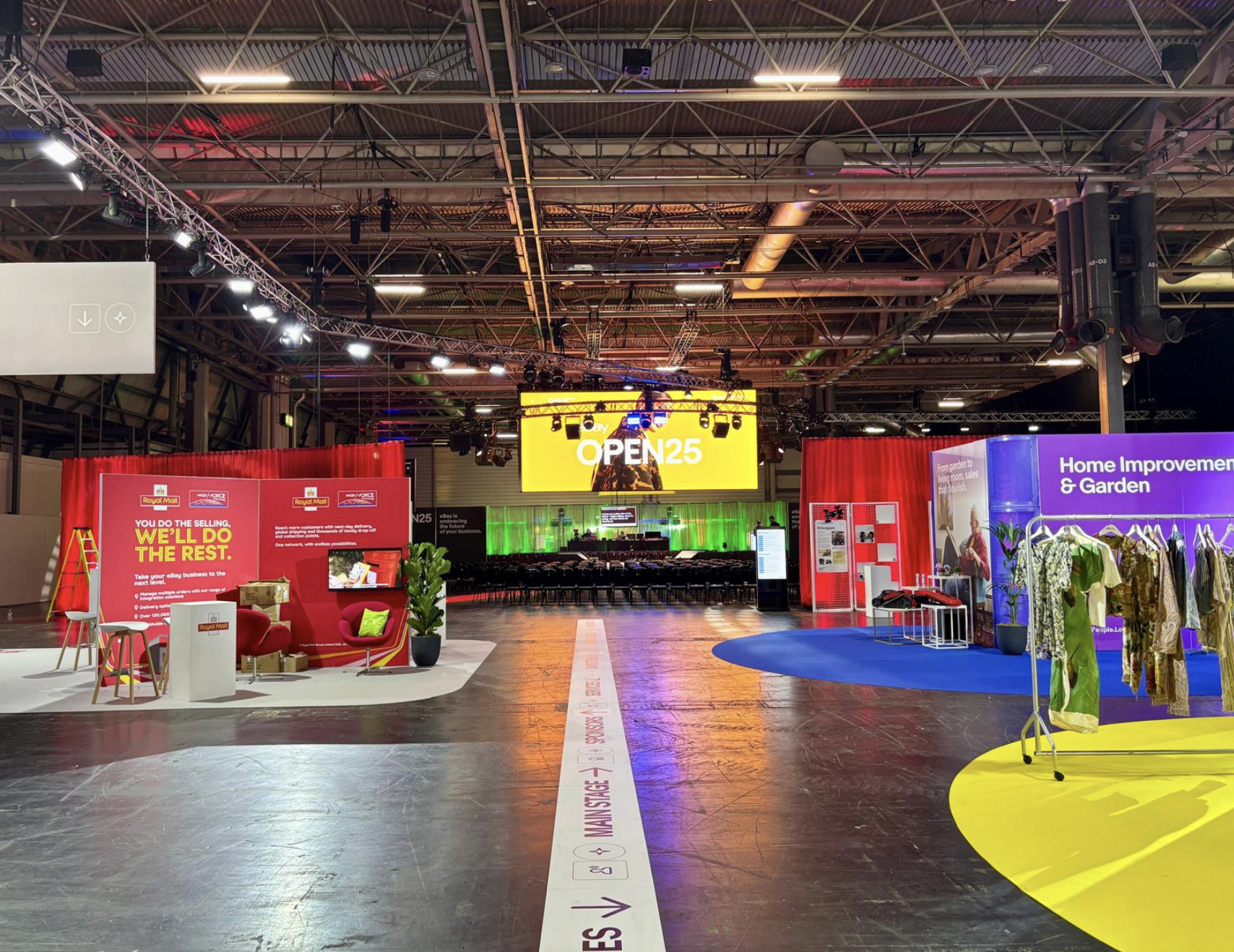 expo hall with various exhibit booths, a stage with a large yellow screen reading 'OPEN25', high industrial ceiling with exposed pipes and lighting rigs.Live show producer, set designer, table scaping, event producer, event consultant, freelance, pri