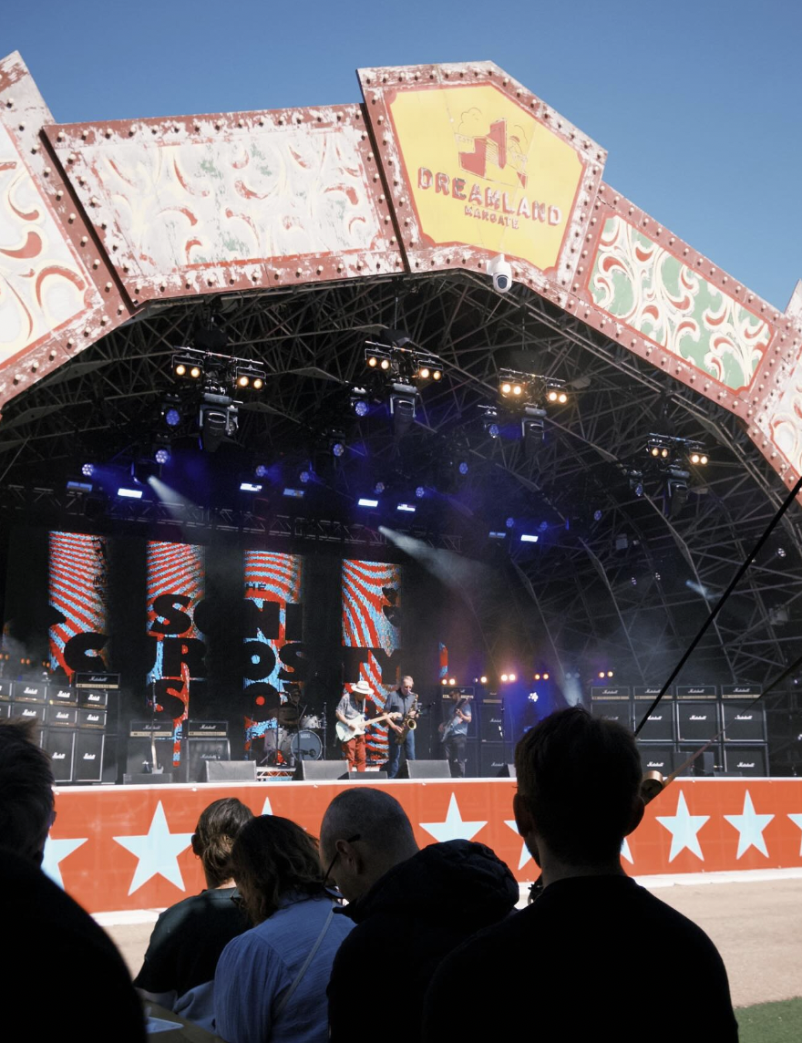 A live outdoor concert at Dreamland Arcade, featuring a band performing on stage with multiple amplifiers and lighting. Audience members are seated and watching the performance under a decorated stage canopy. marshall, jenny bird