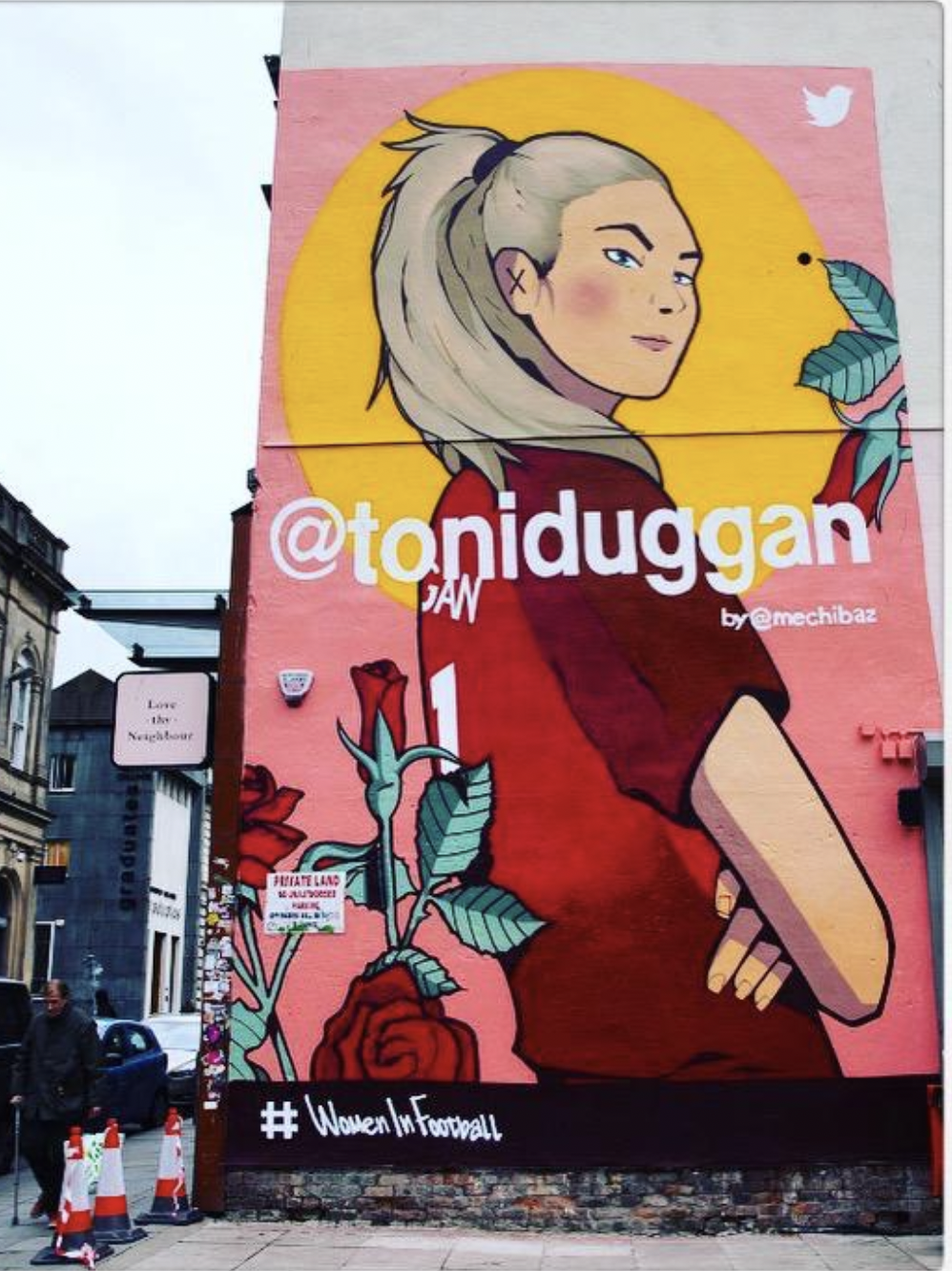 A large mural on a building featuring a stylized drawing of a woman with long blonde hair, wearing a red shirt. The mural includes floral elements like roses and green leaves, and a small bird. The mural has the @toniduggan handle and the hashtag #WomenInFootball.