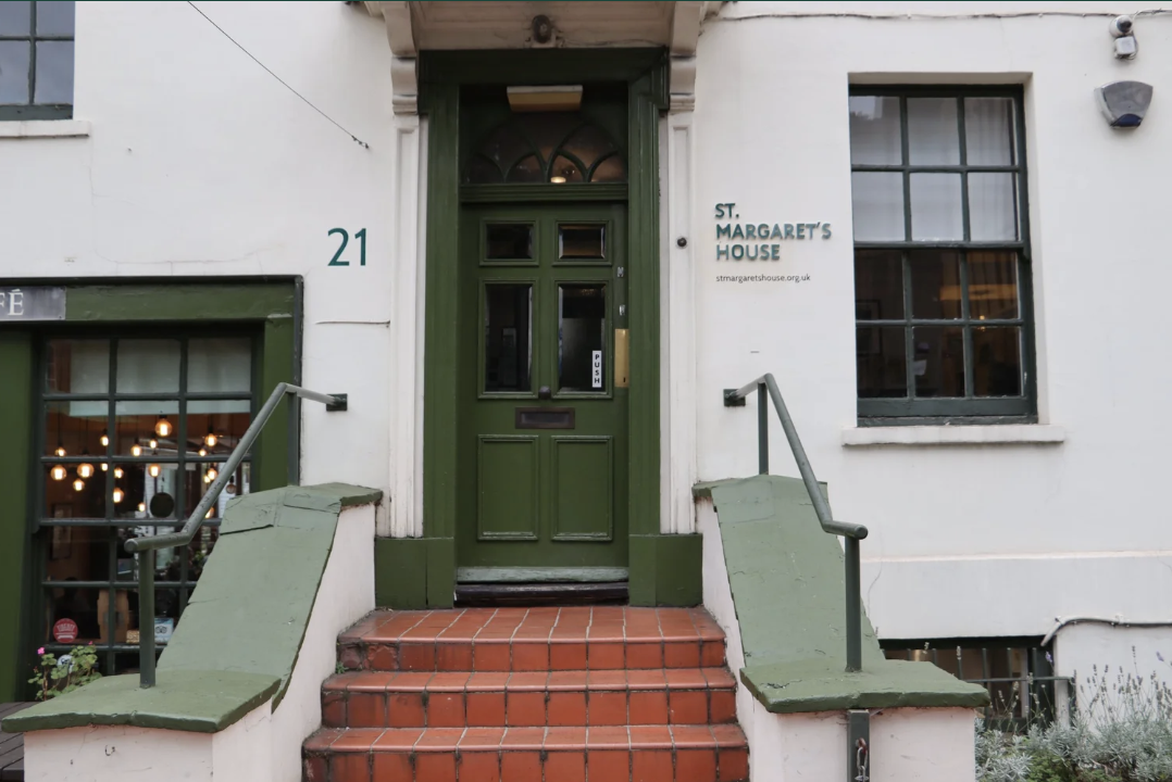 The entrance to St. Margaret's House, a building with a green door and steps, with the street number 21 displayed. There is a sign next to the door indicating the name of the house and a website link.