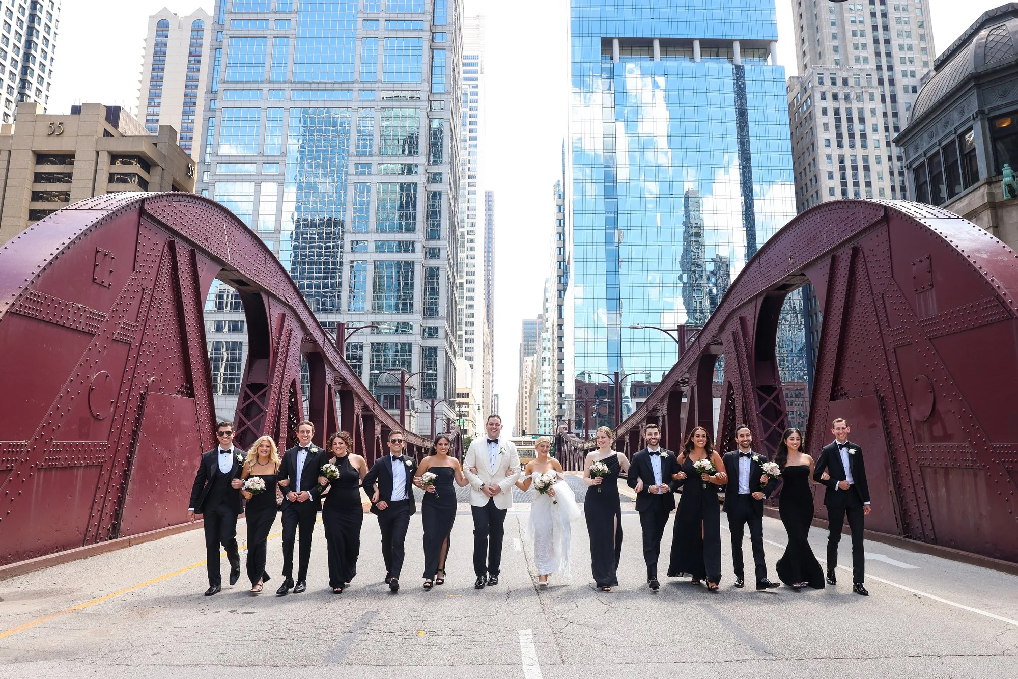 Bridal party of mixed gender walking on city street under a red bridge, surrounded by tall modern glass buildings.