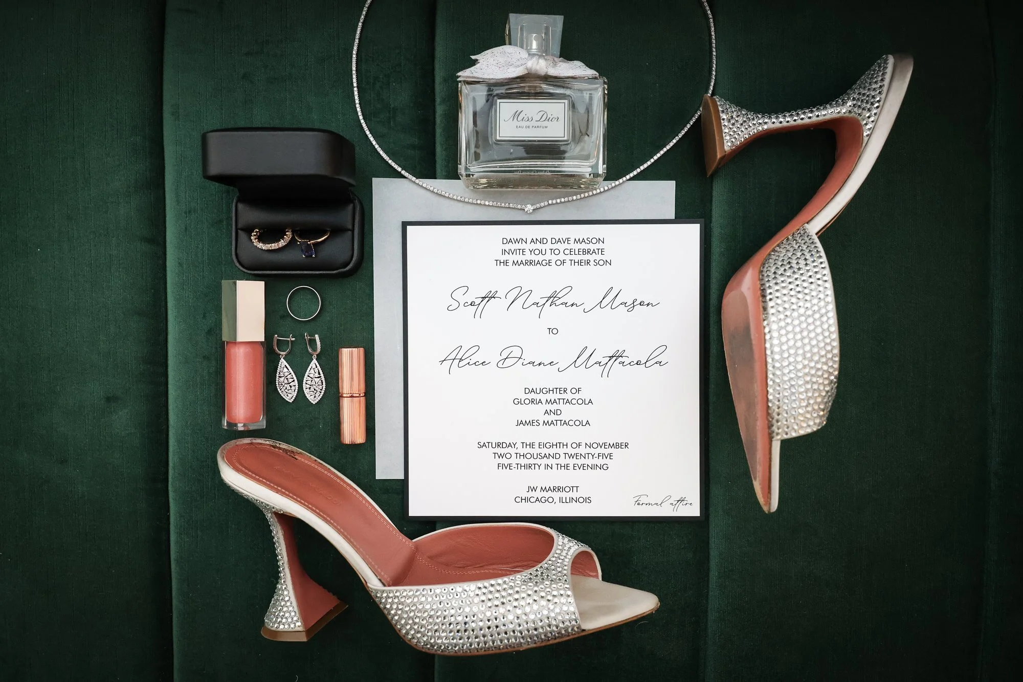 Wedding invitation surrounded by jewelry, shoes, perfume, and makeup on a dark green surface.