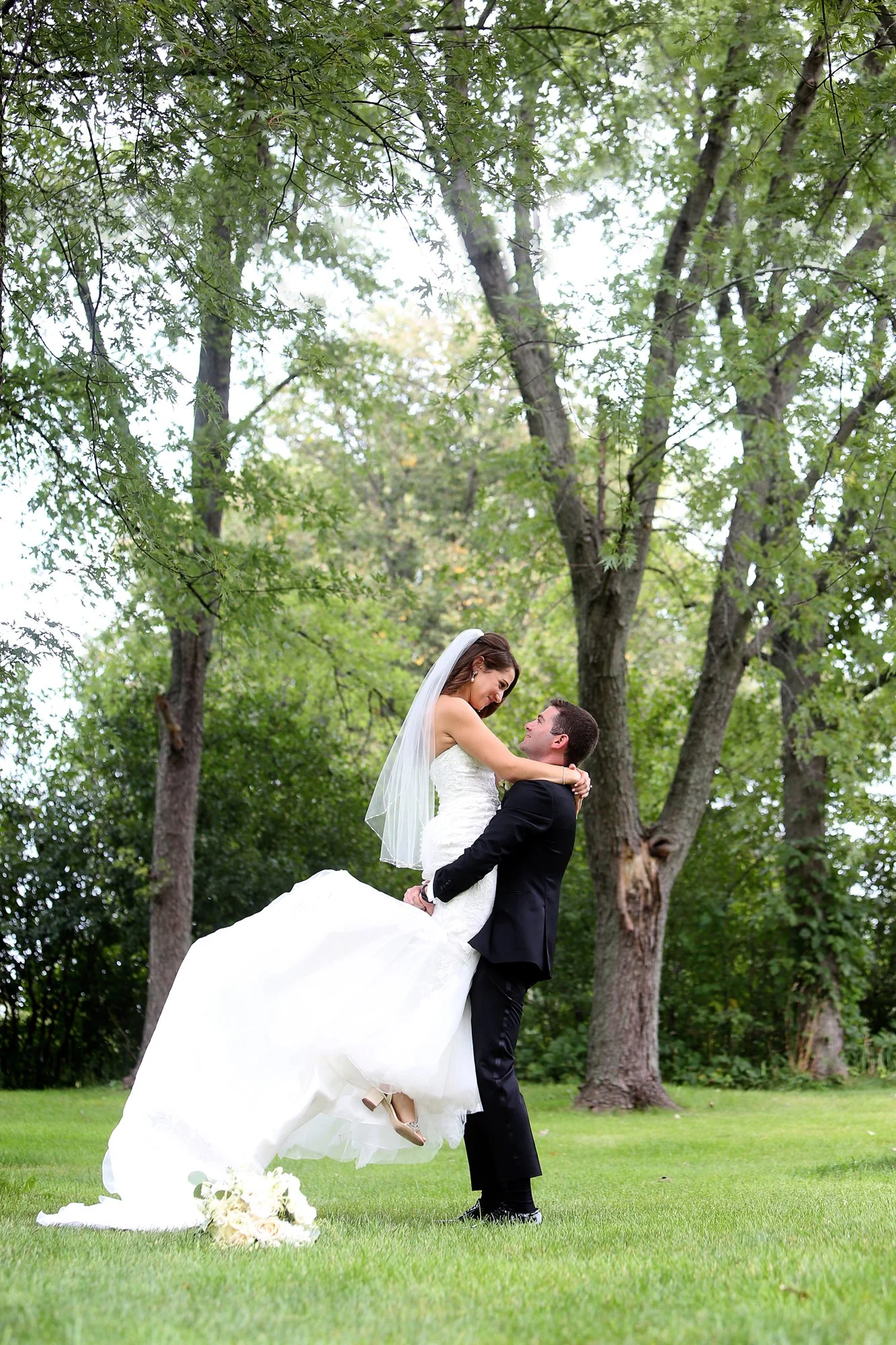 A bride and groom are outdoors in a park with trees, where the groom is lifting the bride, who is wearing a white wedding dress and veil, and the groom is dressed in a black suit.